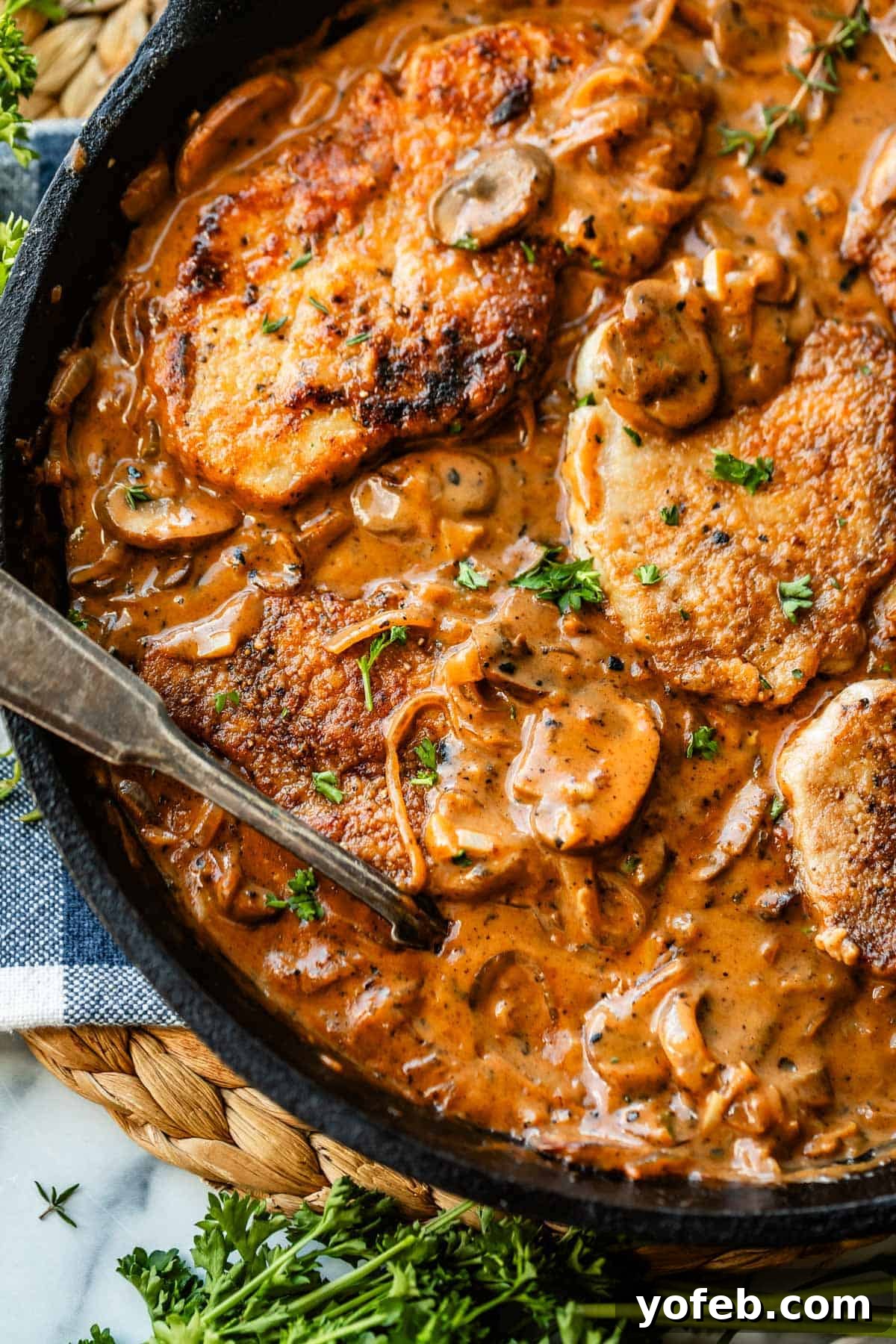 Tender Smothered Pork Chops 4 A skillet filled with golden-brown pork chops in a creamy mushroom sauce, garnished with chopped parsley. A spoon rests in the skillet, and fresh herbs are visible nearby.