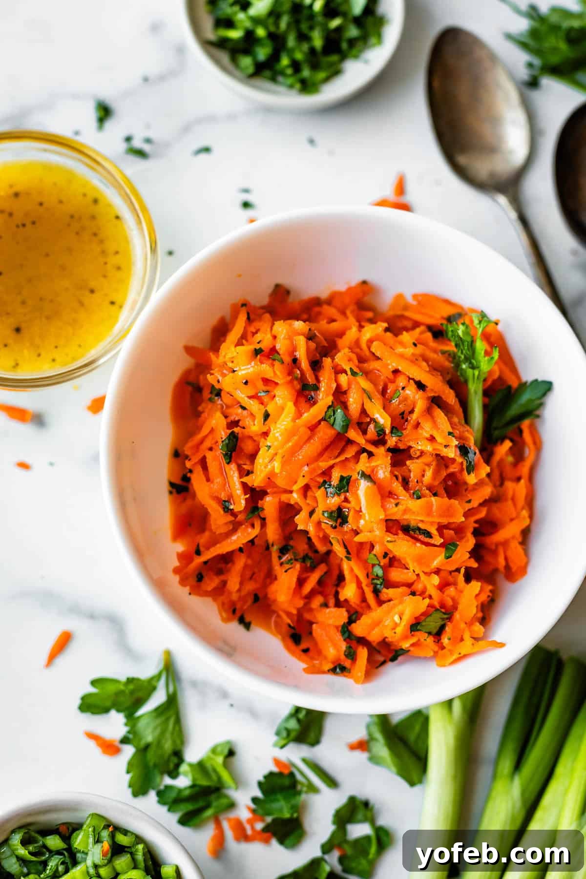 Parisian Carrot Salad 15 A white bowl filled with shredded carrot salad garnished with parsley, surrounded by a small bowl of dressing, a spoon, and chopped herbs on a white surface.
