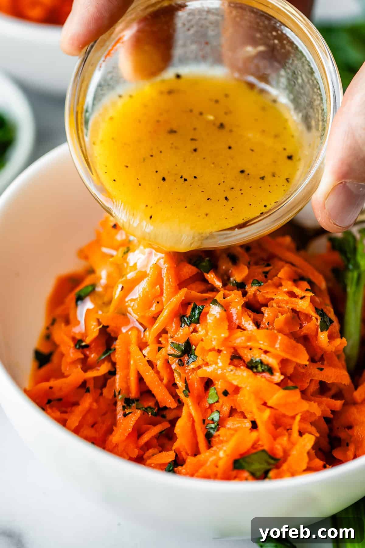 Parisian Carrot Salad 16 A hand pours a small bowl of vinaigrette dressing over a bowl of shredded carrots mixed with fresh herbs.