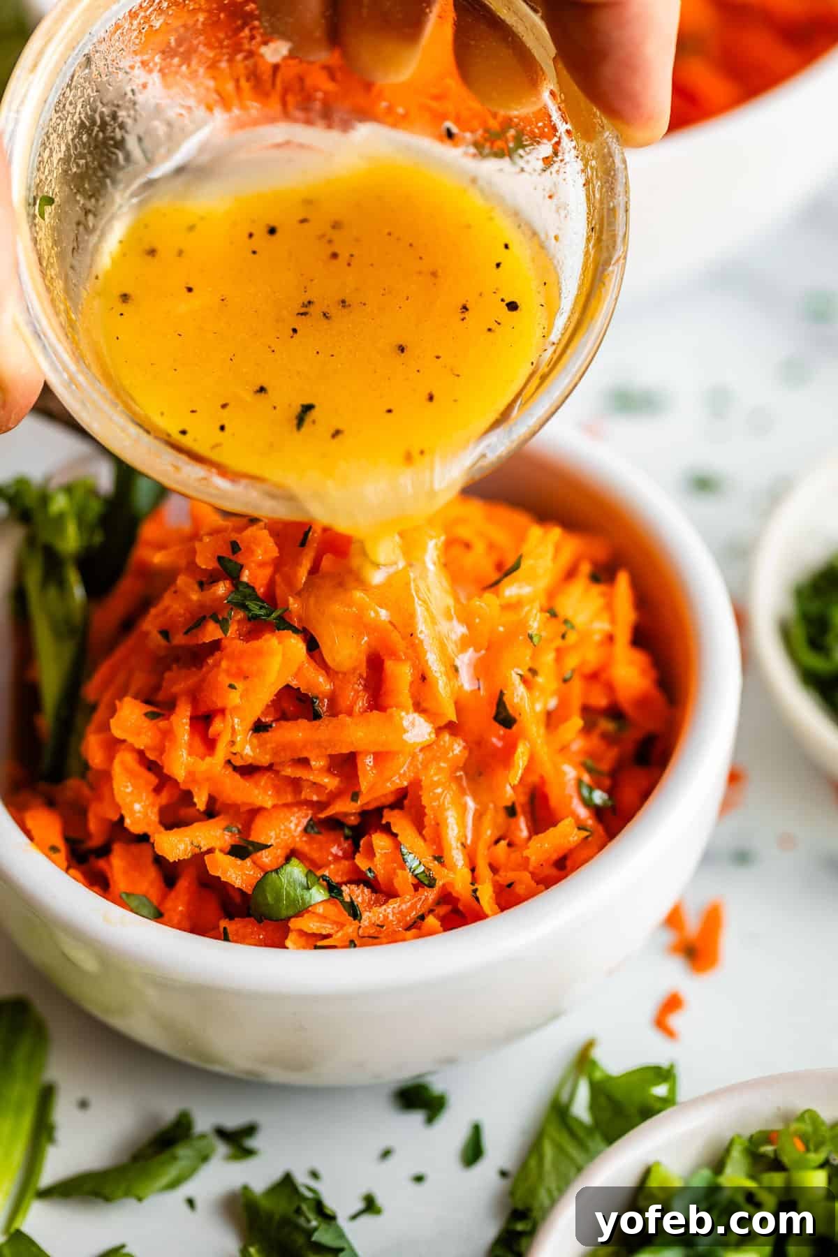Parisian Carrot Salad 3 A hand pours a small bowl of vinaigrette dressing over a serving of shredded carrot salad garnished with chopped herbs, in a white bowl on a white surface with herbs scattered around.