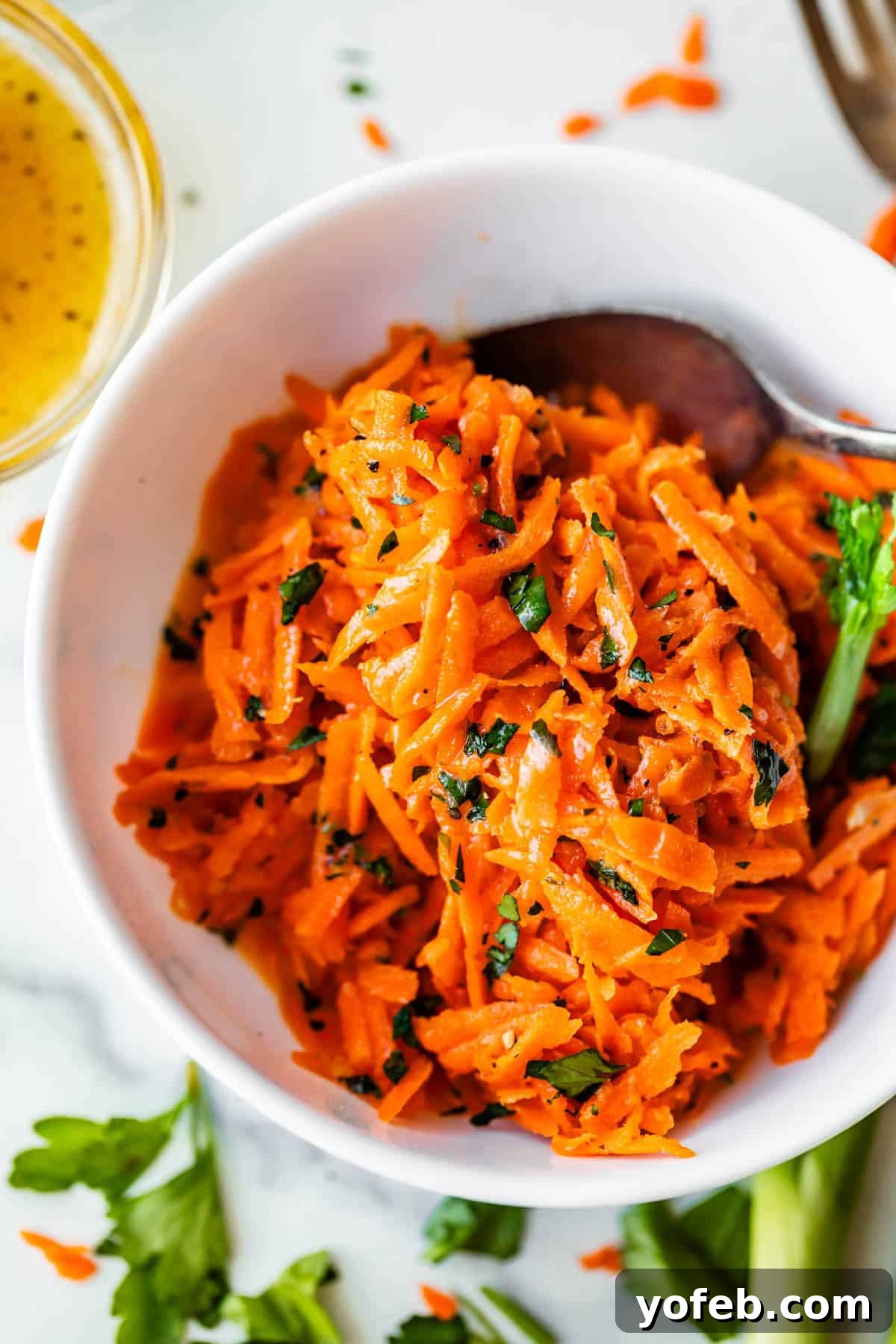 Parisian Carrot Salad 4 A white bowl filled with shredded carrot salad garnished with chopped herbs, with a spoon inside the bowl. Fresh parsley and a small glass container of dressing are nearby on the table.