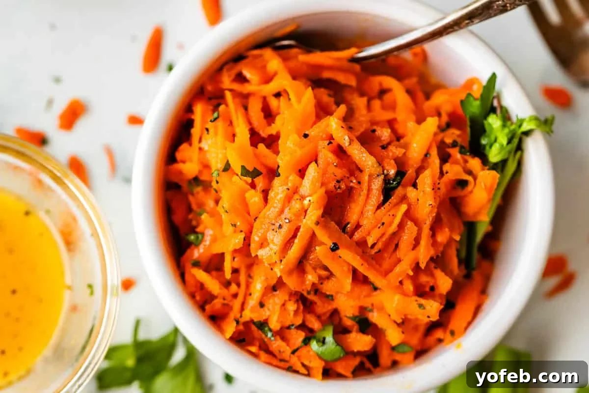 Parisian Carrot Salad 5 A white bowl filled with shredded carrot salad garnished with herbs and cracked black pepper, with a spoon in the bowl. Scattered carrot shreds and a small bowl of yellow dressing are nearby.