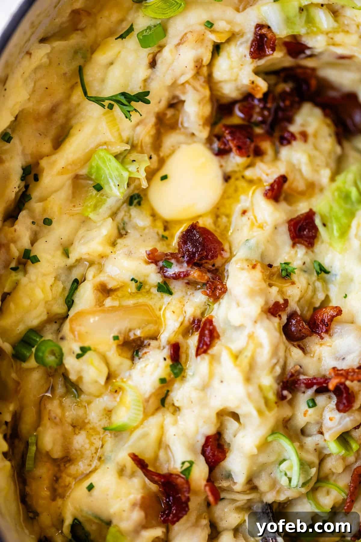 Close-up of creamy mashed potatoes topped with melted butter, chopped green onions, crispy bacon bits, herbs, and visible roasted garlic cloves.