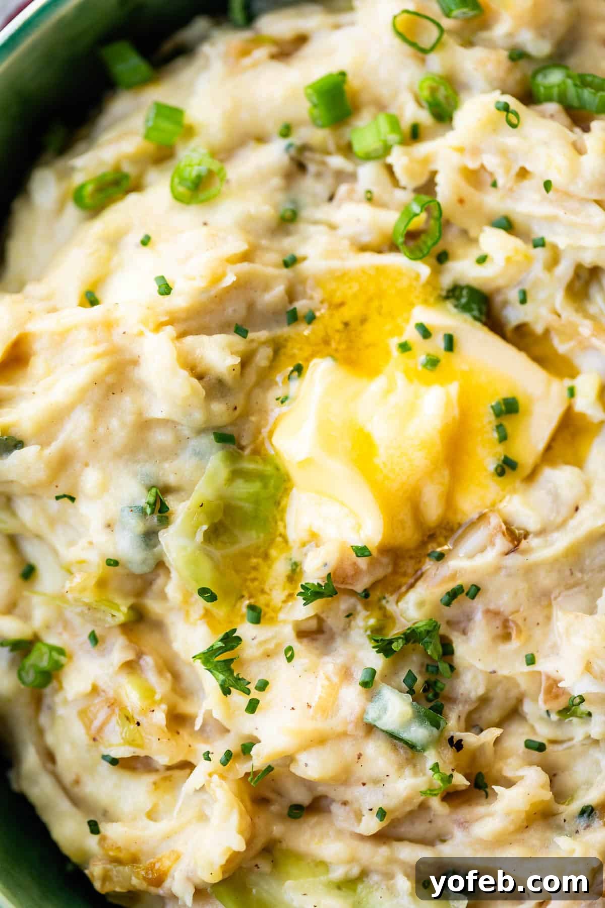 Close-up of creamy mashed potatoes with green onions, topped with a pat of melted butter and sprinkled with chopped chives.