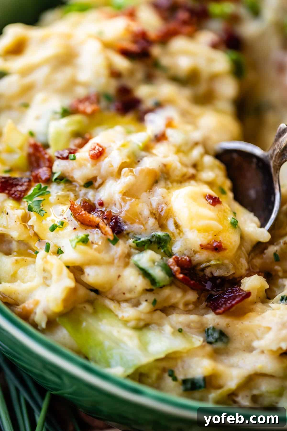 Close-up of a creamy casserole topped with melted cheese, crispy bacon bits, and chopped herbs, with a spoon scooping out a portion from a green dish.