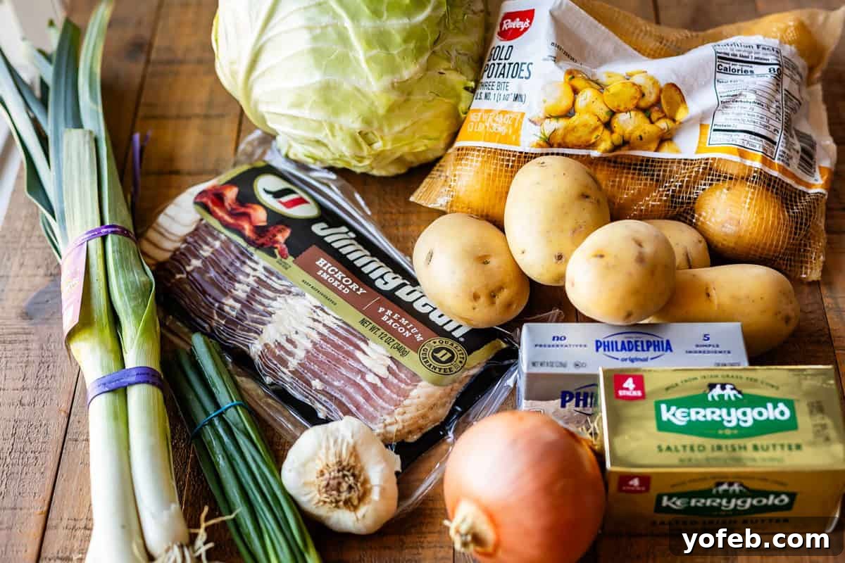 Assorted ingredients on a wooden table, including leeks, a head of cabbage, Yukon Gold potatoes, a yellow onion, garlic, a bundle of scallions, sliced bacon, two sticks of Kerrygold butter, and a package of cream cheese.