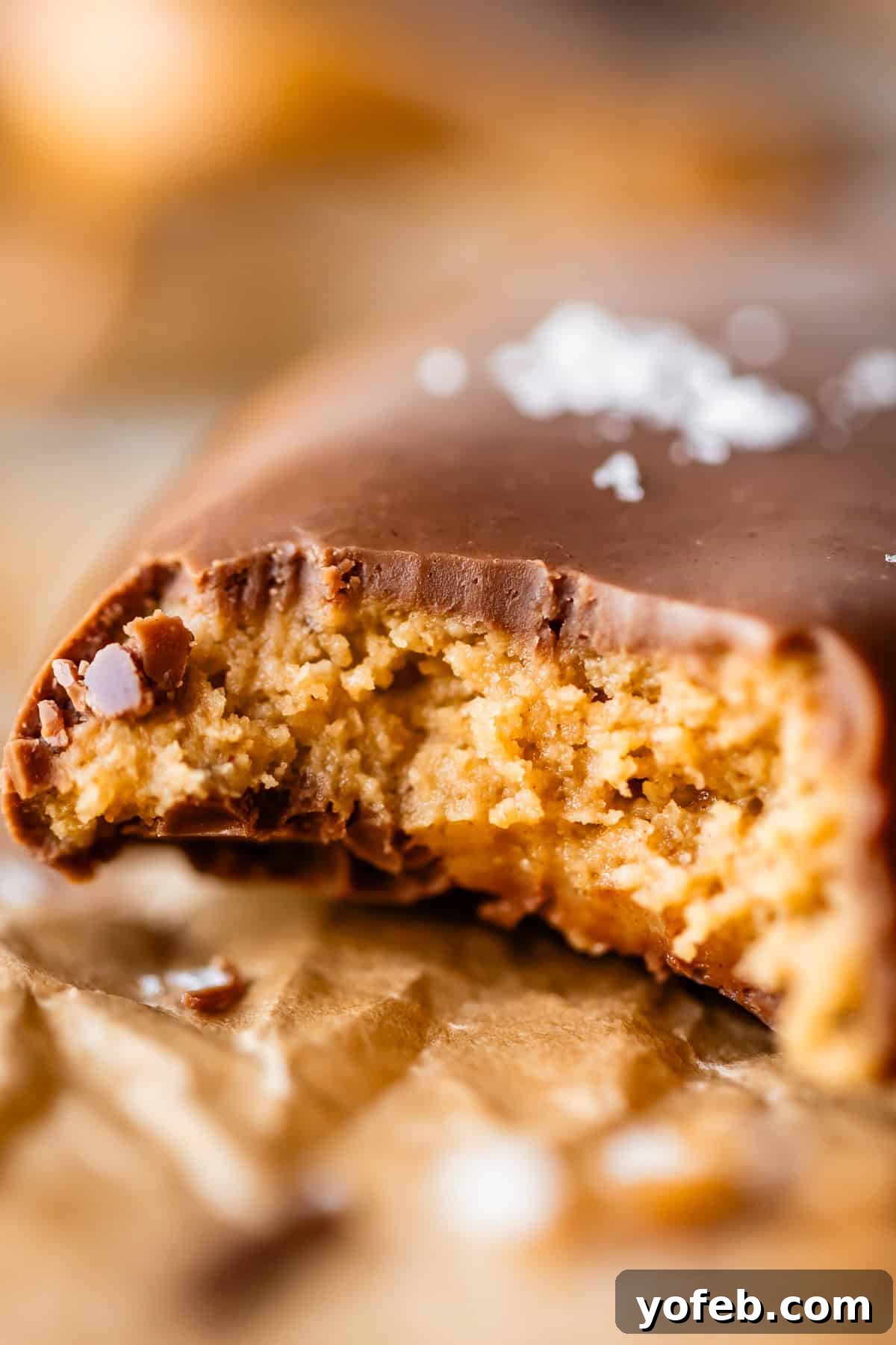 Reese's Easter Peanut Butter Bliss 3 A close-up of a chocolate-covered peanut butter egg with a bite taken out, revealing a creamy peanut butter filling. The bar rests on crinkled brown parchment paper with a sprinkle of sea salt on top.
