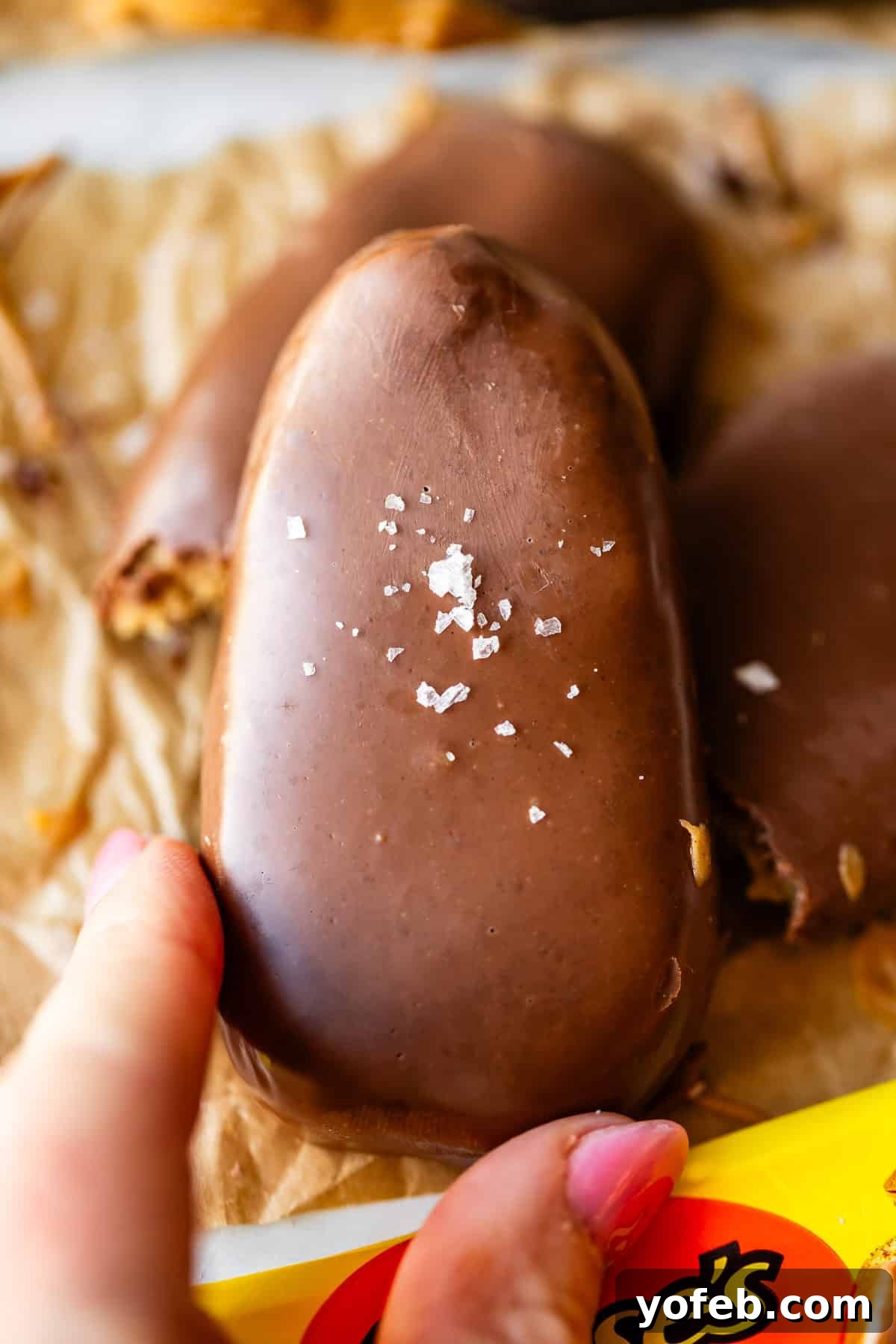 Reese's Easter Peanut Butter Bliss 22 A hand holds a chocolate-covered peanut butter egg topped with flaky sea salt, resting on brown parchment paper with a partially visible Reese’s wrapper below.