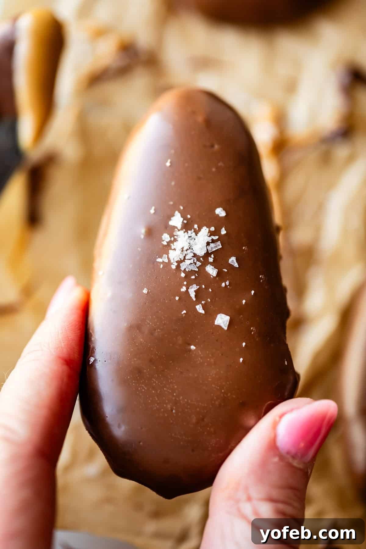 Reese's Easter Peanut Butter Bliss 25 A hand with pink nail polish holds a chocolate-covered peanut butter egg topped with a sprinkle of coarse sea salt, against a blurred background of parchment paper and other treats.