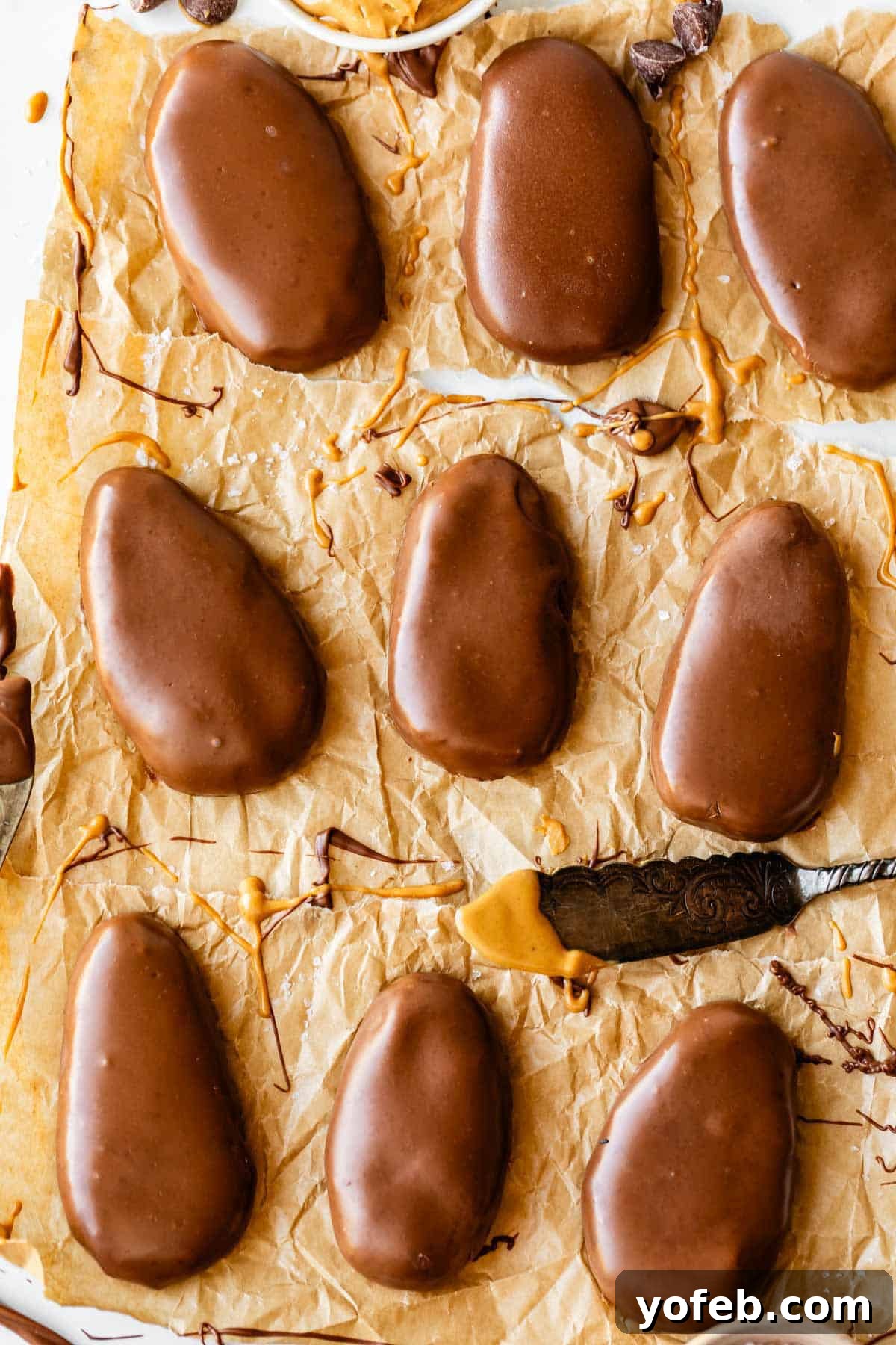 Reese's Easter Peanut Butter Bliss 5 Nine oval-shaped chocolate-covered peanut butter eggs are arranged on crumpled parchment paper, with some chocolate and peanut butter smudges. A small knife with peanut butter rests nearby.
