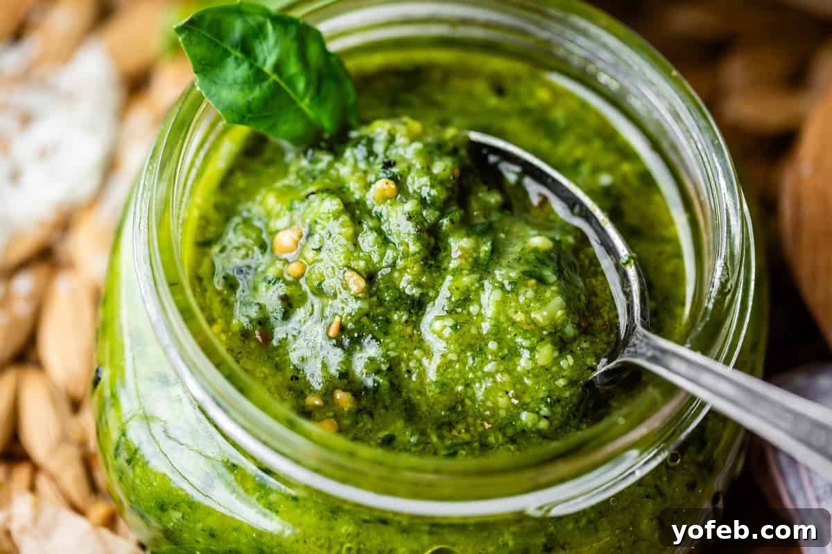Vibrant Basil Pesto 19 A close-up of a glass jar filled with green pesto sauce. A spoon is inside the jar, and a fresh basil leaf garnishes the top. The pesto appears chunky, with visible seeds and herbs.
