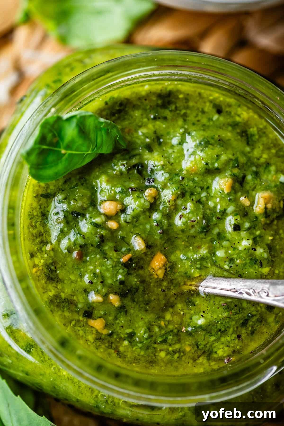Vibrant Basil Pesto 20 Close-up of a jar filled with vibrant green pesto sauce, garnished with a fresh basil leaf. A spoon is partially dipped in the textured sauce, showing bits of nuts and herbs throughout.