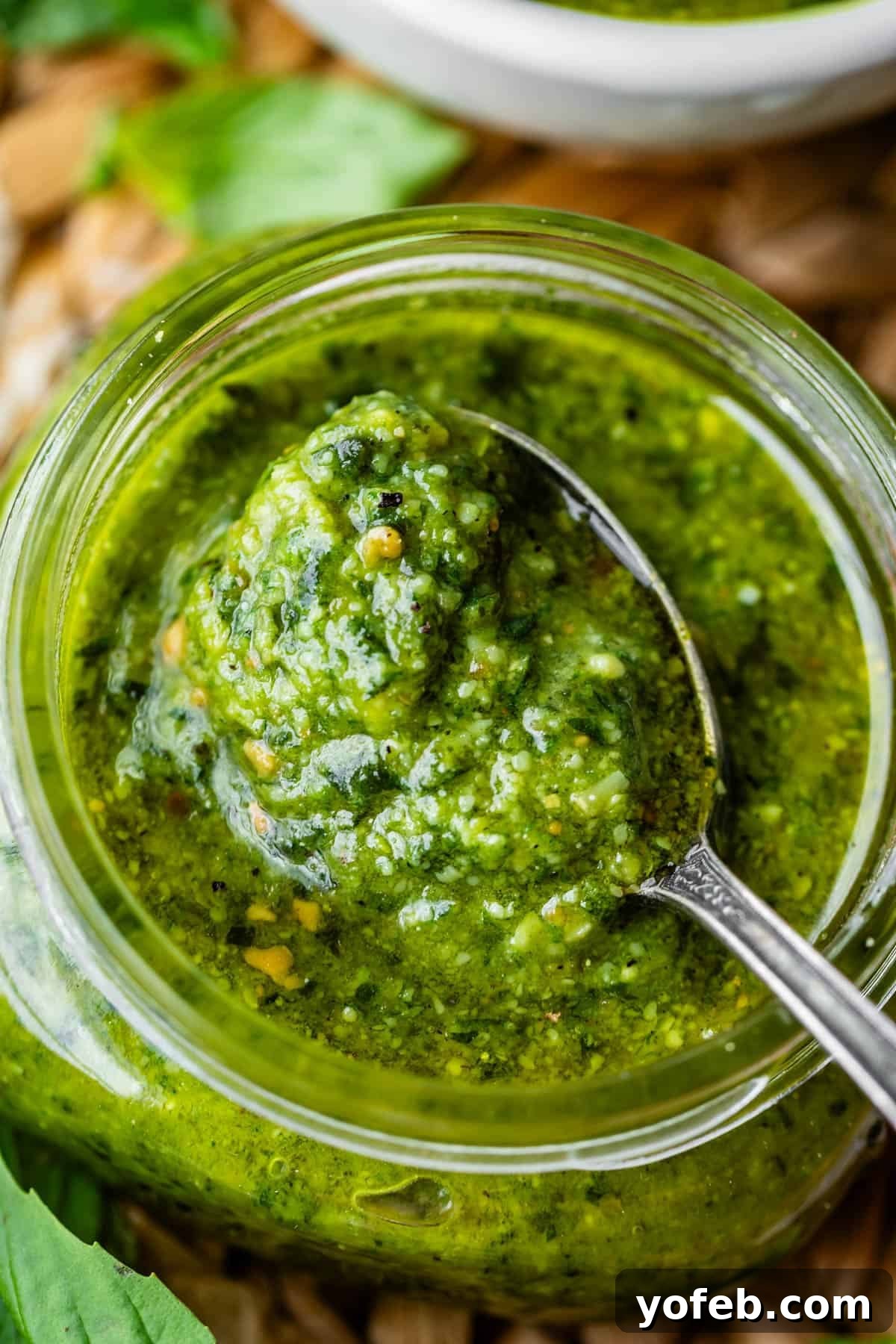 Vibrant Basil Pesto 4 Close-up of a jar of vibrant green pesto sauce with a spoonful being lifted from the jar; fresh herbs are visible in the background.