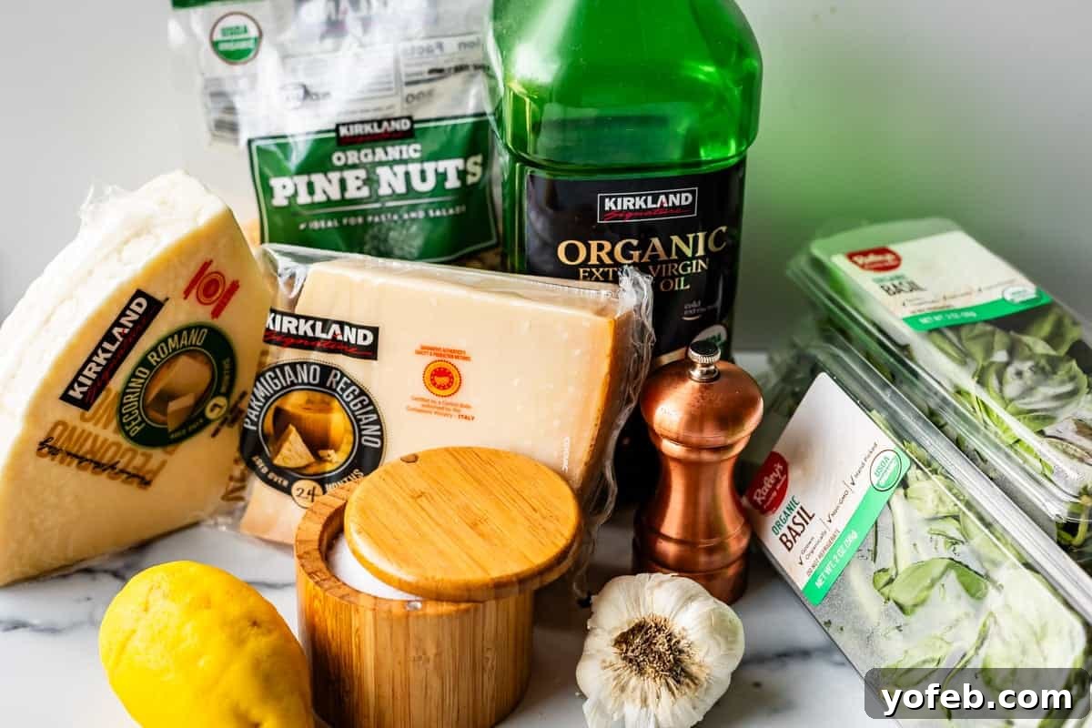 Vibrant Basil Pesto 6 A collection of ingredients for pesto, including parmesan and romano cheese wedges, organic pine nuts, extra virgin olive oil, fresh basil, a lemon, garlic bulb, salt cellar, and a wooden pepper grinder on a marble surface.