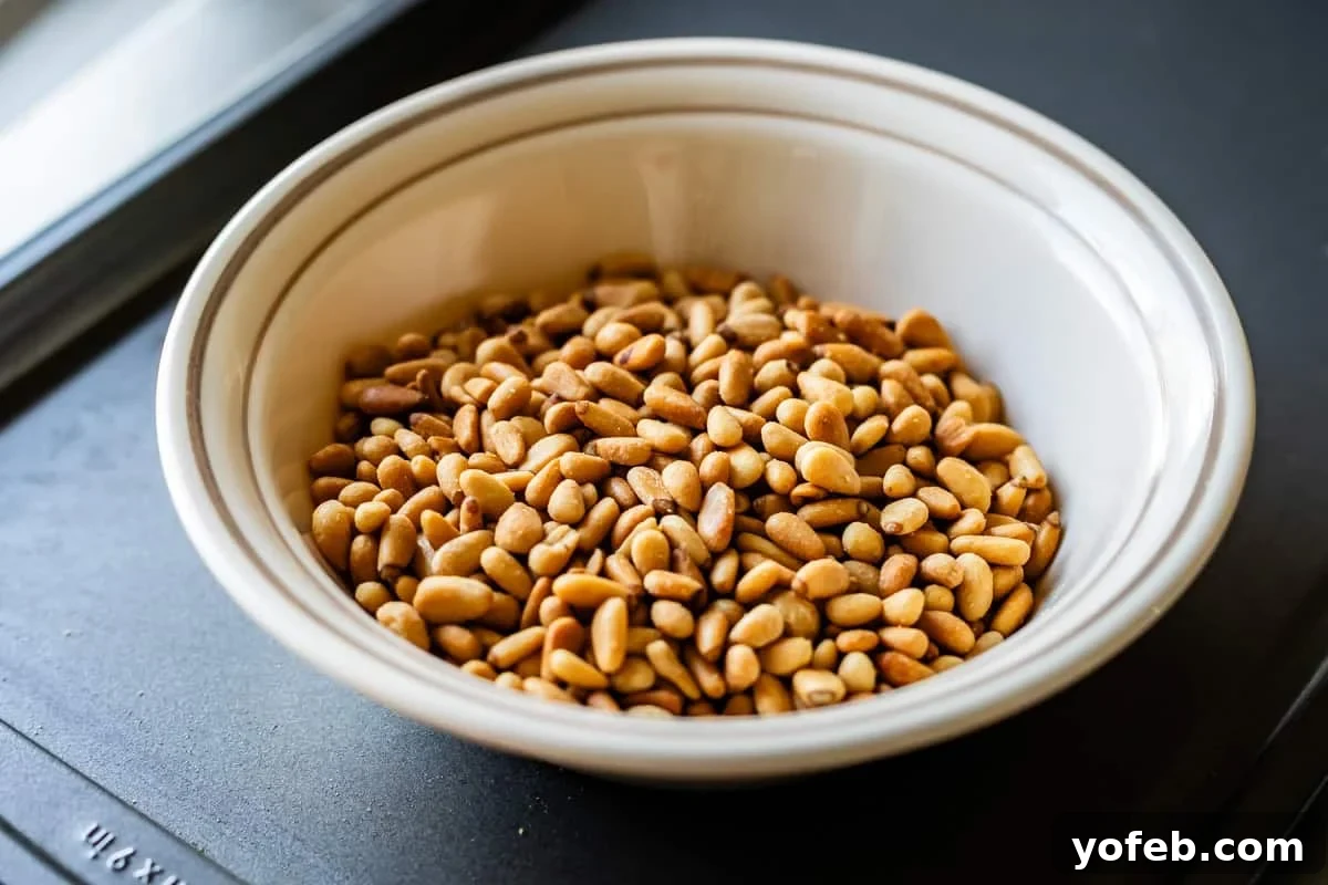 Vibrant Basil Pesto 8 A white bowl filled with toasted pine nuts sits on a dark surface.
