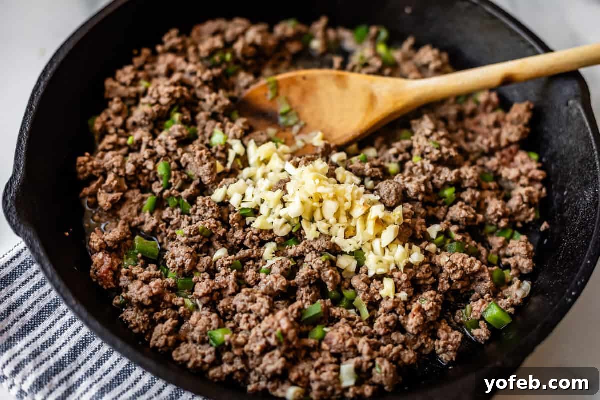 Crispy Egg Roll Bowl: Stir-Fry Recipe Without Wrappers 11 Ground beef is being cooked in a black skillet with chopped green onions and minced garlic, stirred with a wooden spoon. The skillet rests on a striped cloth.