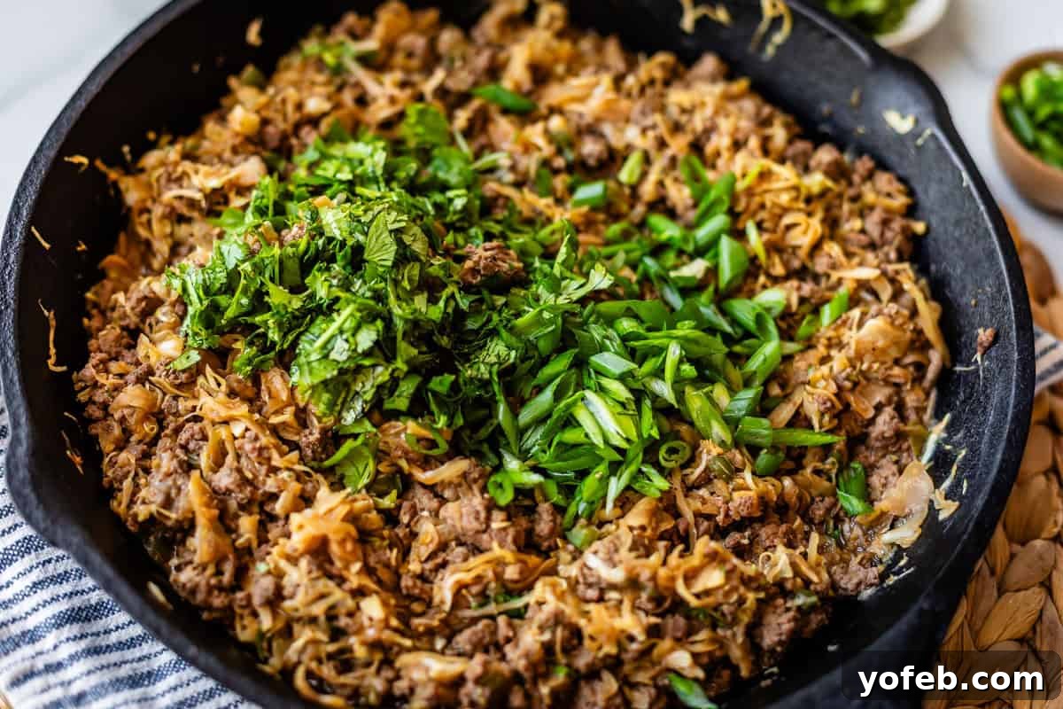 Crispy Egg Roll Bowl: Stir-Fry Recipe Without Wrappers 15 Ground beef, shredded cabbage, and onions cooked in a black skillet, topped with chopped fresh green onions and herbs. The skillet sits on a woven mat beside a striped towel.