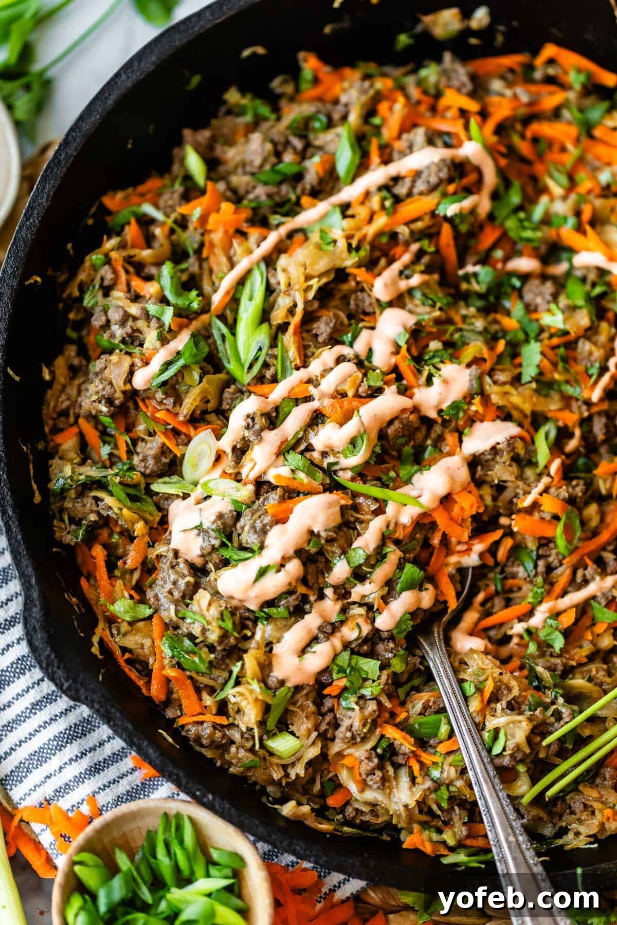 Crispy Egg Roll Bowl: Stir-Fry Recipe Without Wrappers 3 A skillet filled with a colorful mixture of shredded vegetables and ground meat, topped with chopped green onions, cilantro, and a drizzle of creamy sauce. A spoon rests in the skillet for serving.