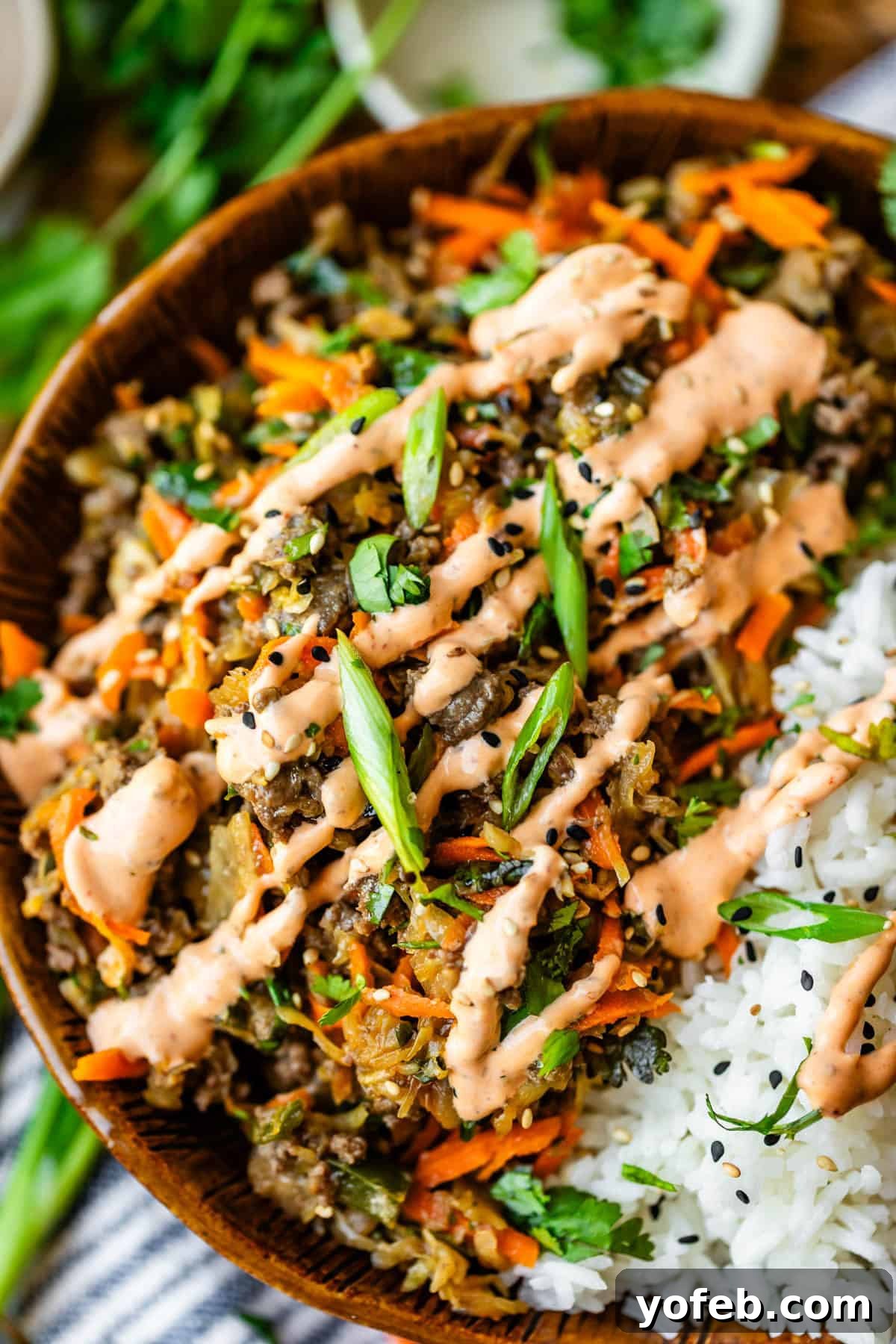 Crispy Egg Roll Bowl: Stir-Fry Recipe Without Wrappers 19 A close-up of a bowl filled with white rice and a colorful stir-fry topped with sliced green onions, shredded carrots, black sesame seeds, fresh herbs, and a drizzle of creamy orange sauce.