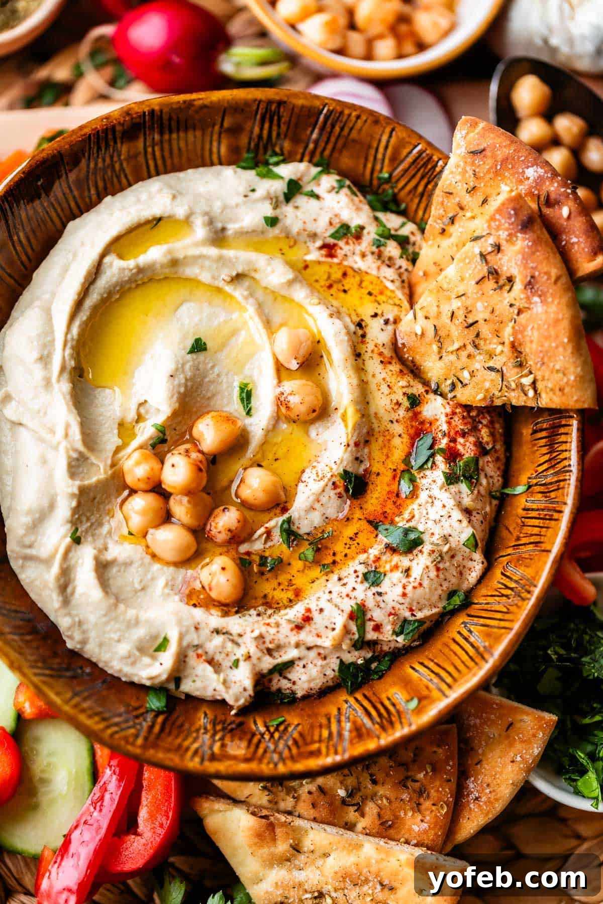 Creamy Homemade Hummus from Scratch: Classic Chickpea Dip Recipe 8 A bowl of creamy hummus topped with whole chickpeas, olive oil, spices, and herbs, served with wedges of pita bread and surrounded by fresh vegetables.