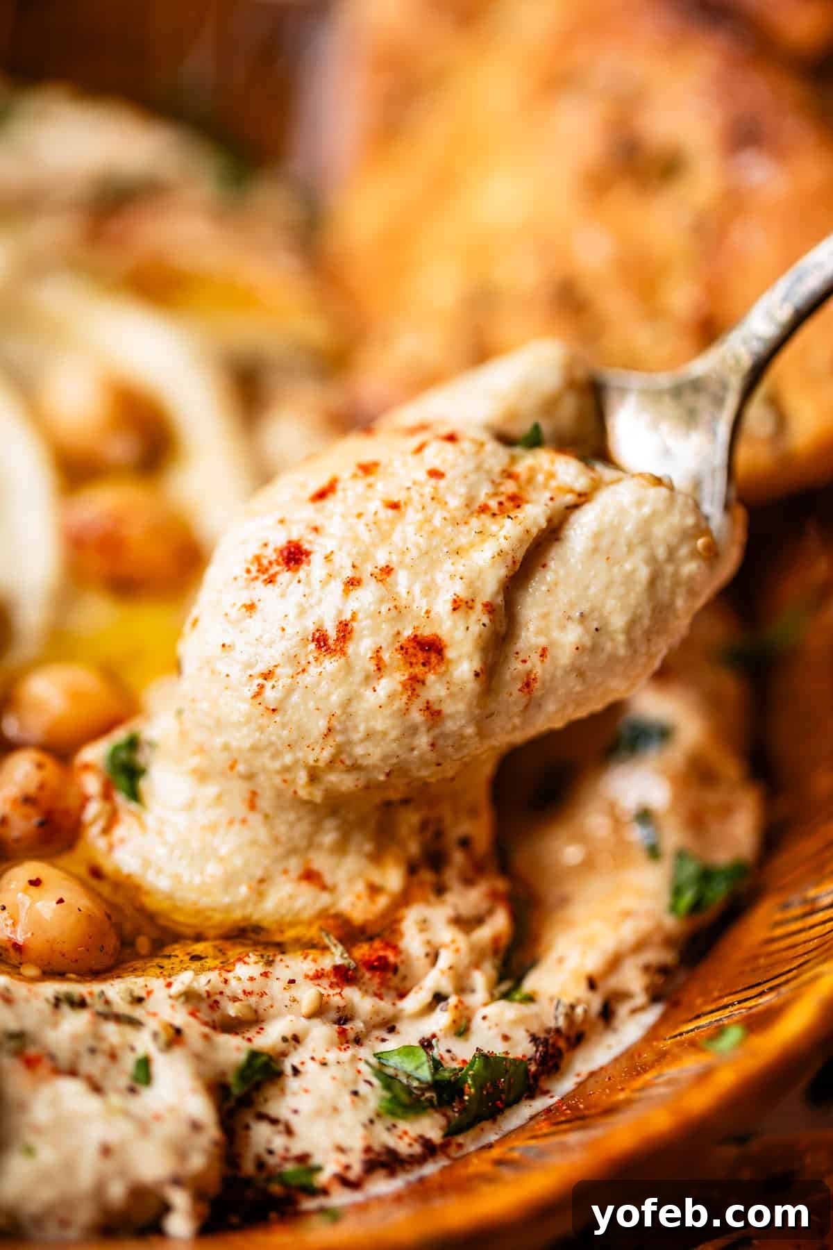 Creamy Homemade Hummus from Scratch: Classic Chickpea Dip Recipe 9 A close-up of a spoon scooping creamy hummus, garnished with chickpeas, chopped herbs, olive oil, and paprika.
