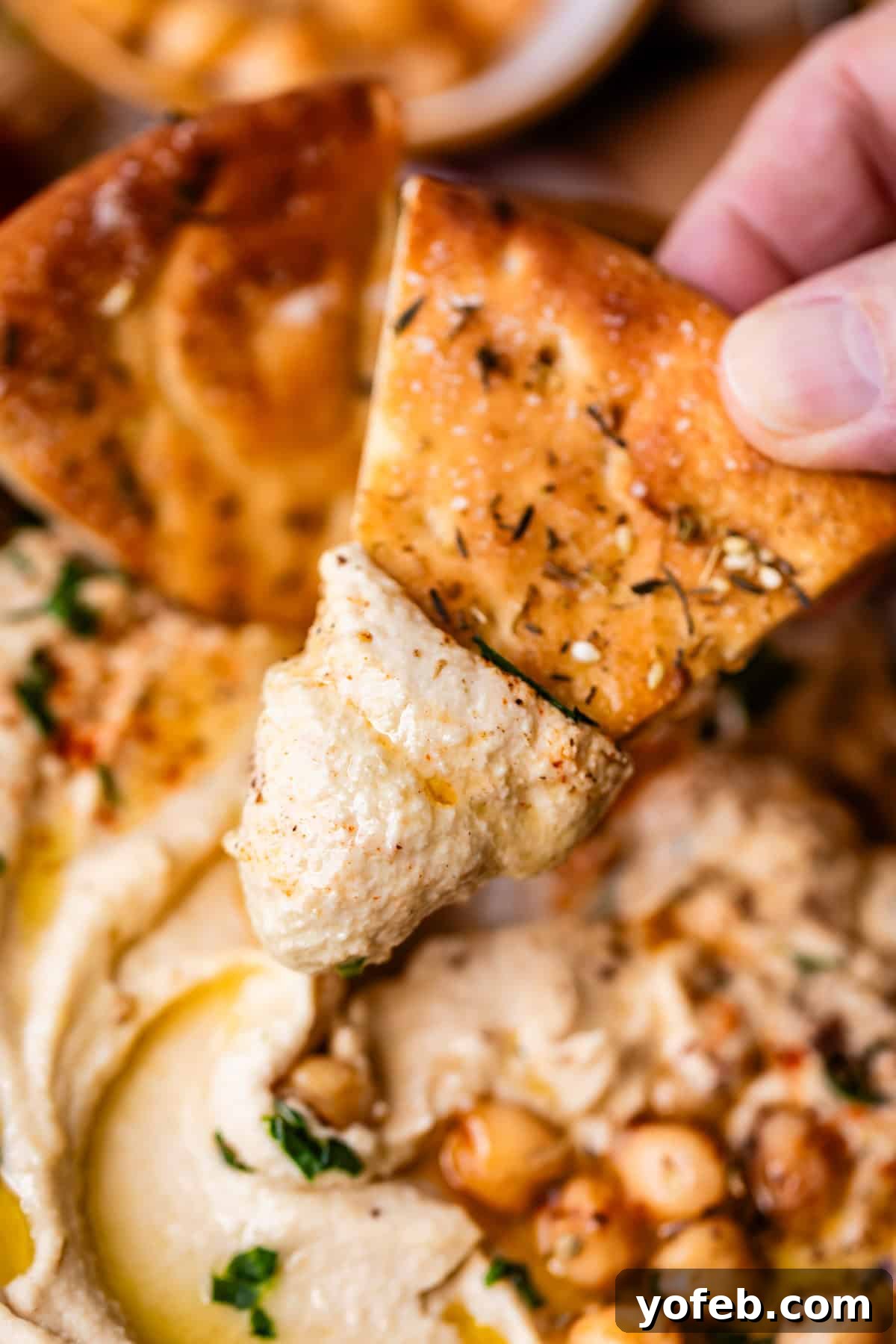 Creamy Homemade Hummus from Scratch: Classic Chickpea Dip Recipe 3 A hand dips a piece of seasoned pita bread into creamy hummus, garnished with whole chickpeas, olive oil, and herbs.