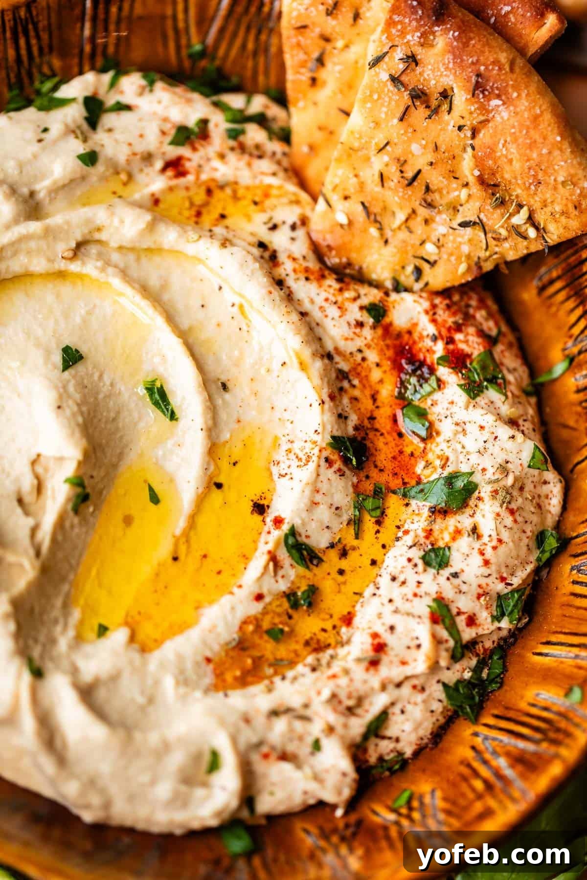 Creamy Homemade Hummus from Scratch: Classic Chickpea Dip Recipe 4 A close-up of creamy hummus drizzled with olive oil, garnished with paprika, chopped parsley, and lemon slices, served with pieces of seasoned pita bread on a rustic brown plate.