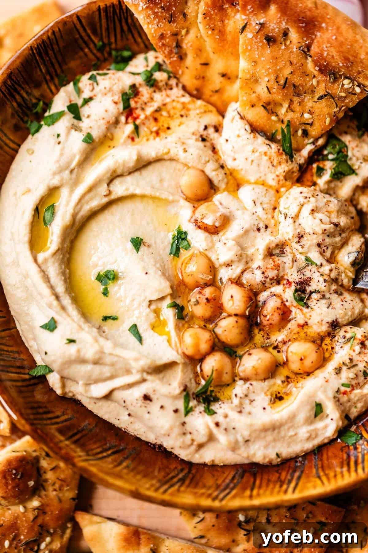 Creamy Homemade Hummus from Scratch: Classic Chickpea Dip Recipe 5 A bowl of creamy hummus topped with whole chickpeas, olive oil, chopped parsley, and spices, served with pieces of seasoned flatbread.