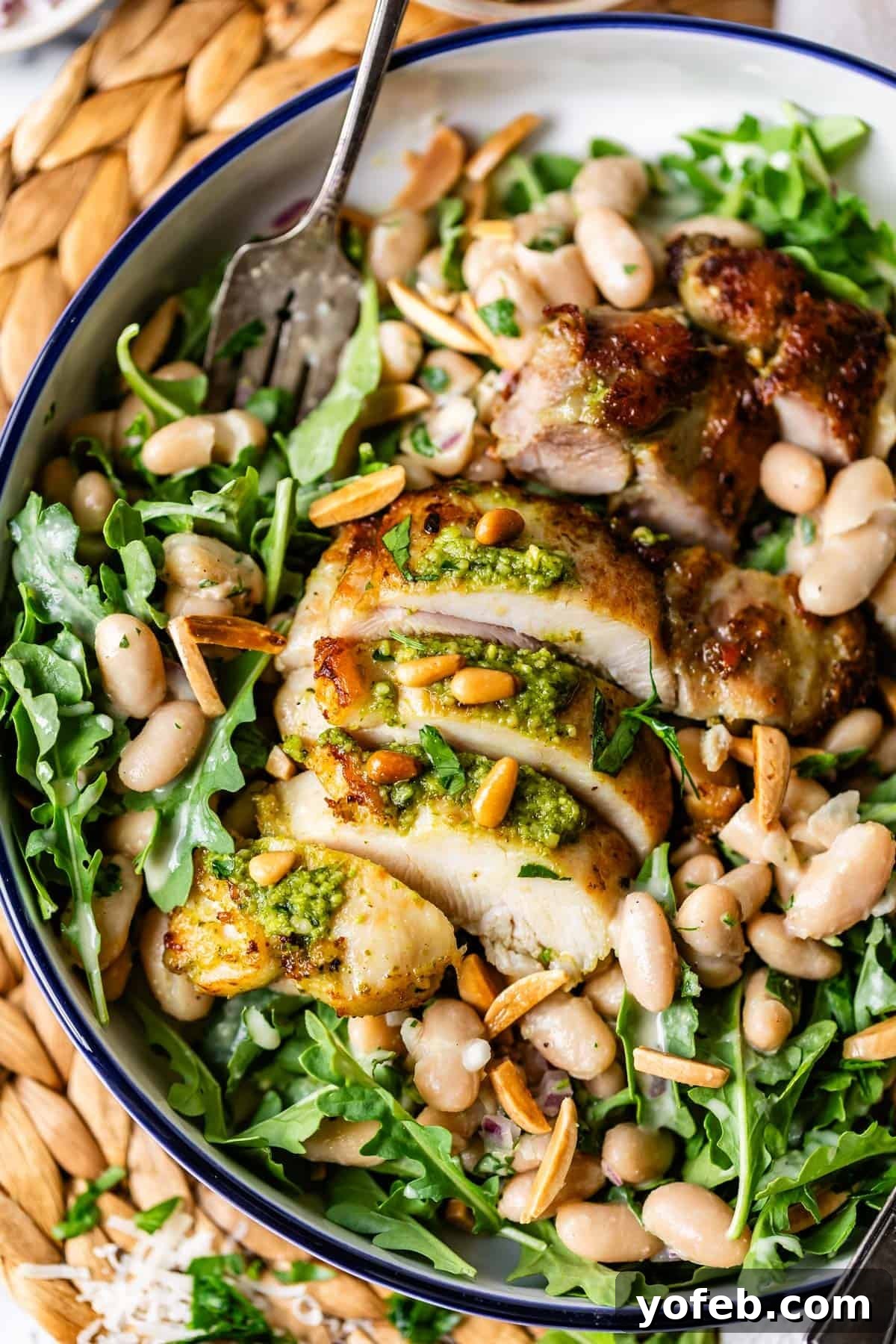 Basil Chicken Salad with Peppery Arugula and Lemon Vinaigrette 2 A bowl filled with sliced grilled chicken topped with pesto, served on a bed of arugula, white beans, toasted pine nuts, and slivered almonds, with a fork resting on the side.