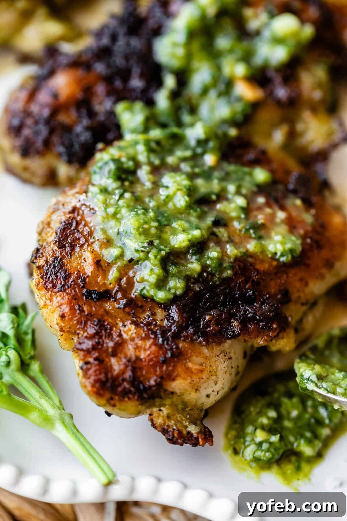 Basil Chicken Salad with Peppery Arugula and Lemon Vinaigrette 8 Close-up of a piece of grilled chicken topped with green pesto sauce, served on a white plate with a small sprig of fresh herbs on the side. The chicken has a crispy, golden-brown skin.