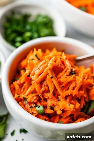 Basil Chicken Salad with Peppery Arugula and Lemon Vinaigrette 11 A white bowl filled with shredded carrot salad garnished with chopped herbs and freshly ground black pepper, with a spoon in the bowl and blurred bowls of greens in the background.