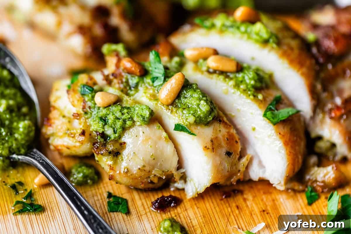 Creamy Pesto Chicken Recipe with Garlic and Parmesan 11 Close-up of sliced grilled chicken breast topped with green pesto sauce, pine nuts, and chopped herbs on a wooden cutting board, with a spoon of pesto beside it.