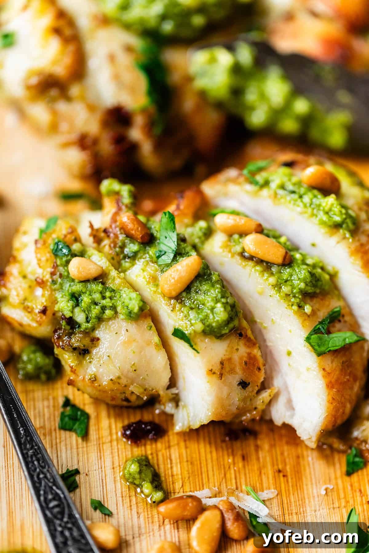 Creamy Pesto Chicken Recipe with Garlic and Parmesan 3 Sliced grilled chicken breast topped with green pesto, pine nuts, and chopped parsley, served on a wooden board.