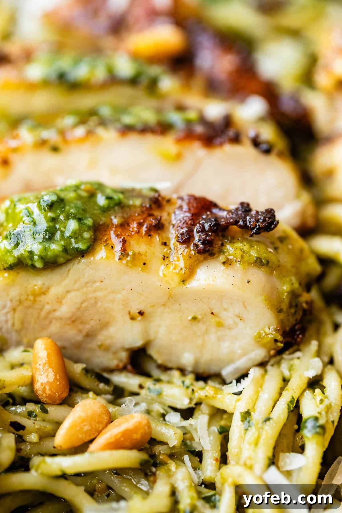 Creamy Pesto Chicken Recipe with Garlic and Parmesan 4 Close-up of sliced, golden-brown chicken breast topped with pesto, resting on a bed of pesto-coated spaghetti with pine nuts and grated cheese.
