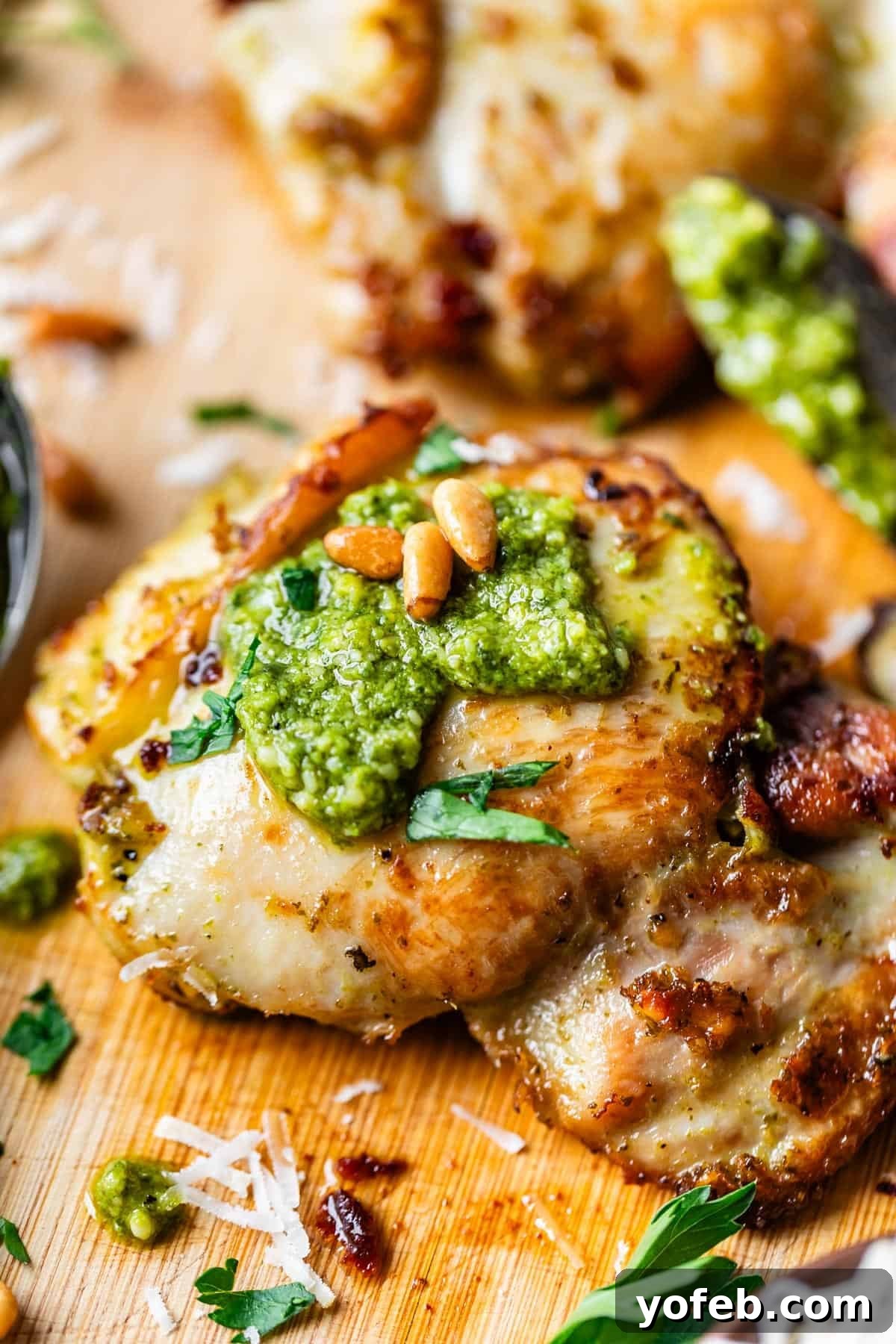 Creamy Pesto Chicken Recipe with Garlic and Parmesan 5 Close-up of a grilled chicken thigh topped with green pesto, pine nuts, and chopped parsley, served on a wooden board with grated cheese and herb garnishes.