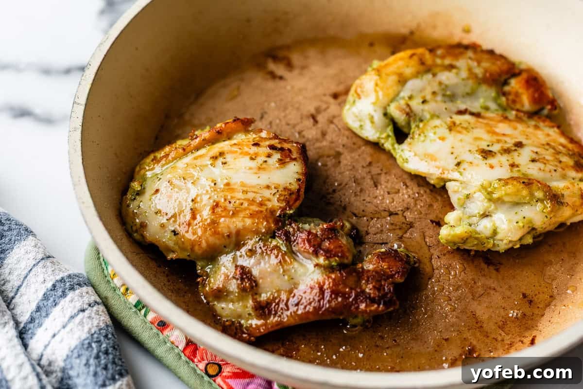 Creamy Pesto Chicken Recipe with Garlic and Parmesan 9 Two browned chicken thighs are cooking in a beige skillet with some oil, resting on a patterned oven mitt. A striped kitchen towel is visible nearby on a marble countertop.