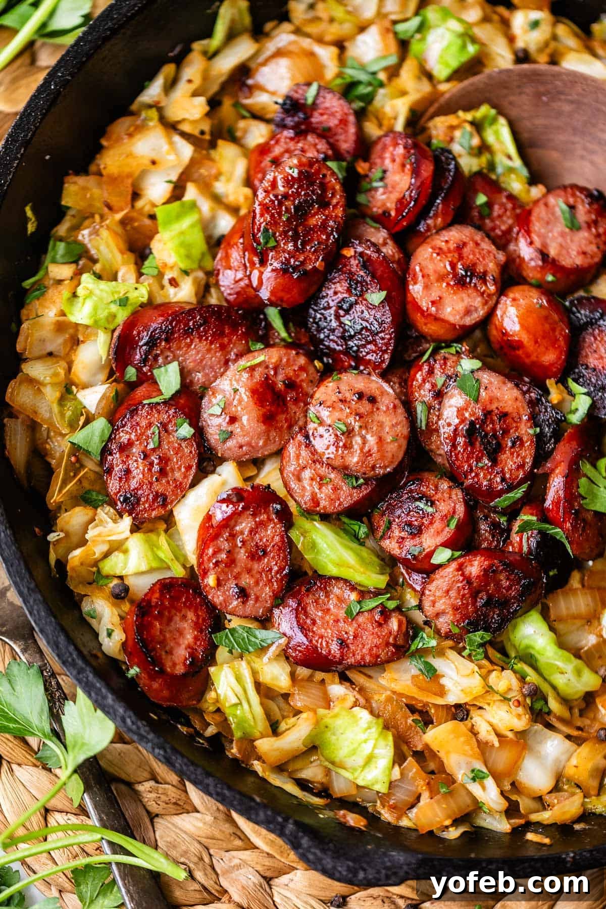 One-Pan Kielbasa and Cabbage Skillet Recipe for Quick Dinners 2 Sliced, browned sausage is served over sautéed cabbage and onions in a black skillet, garnished with fresh parsley. A wooden spoon rests in the pan, and a woven mat is partially visible underneath.