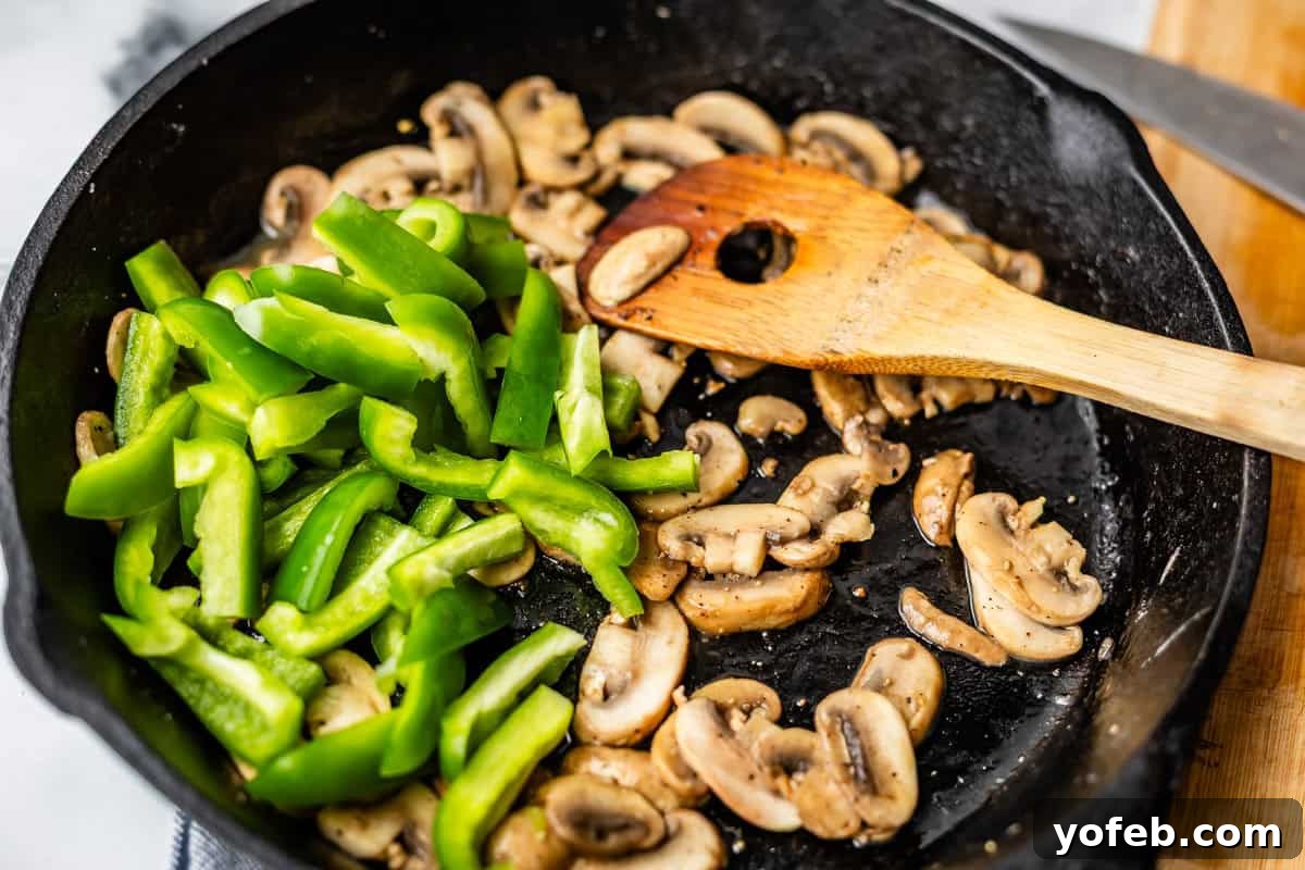 Authentic Philly Cheesesteak Recipe: How to Make It at Home 11 Sliced green bell peppers and mushrooms being sautéed in a black skillet, with a wooden spatula resting inside the pan.