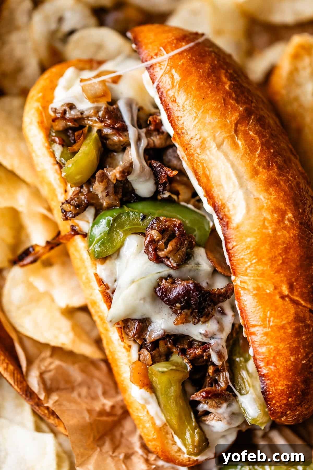 Authentic Philly Cheesesteak Recipe: How to Make It at Home 3 A close-up of a Philly cheesesteak sandwich on a toasted bun, filled with melted cheese, sautéed green bell peppers, onions, and thinly sliced steak, served with potato chips in the background.