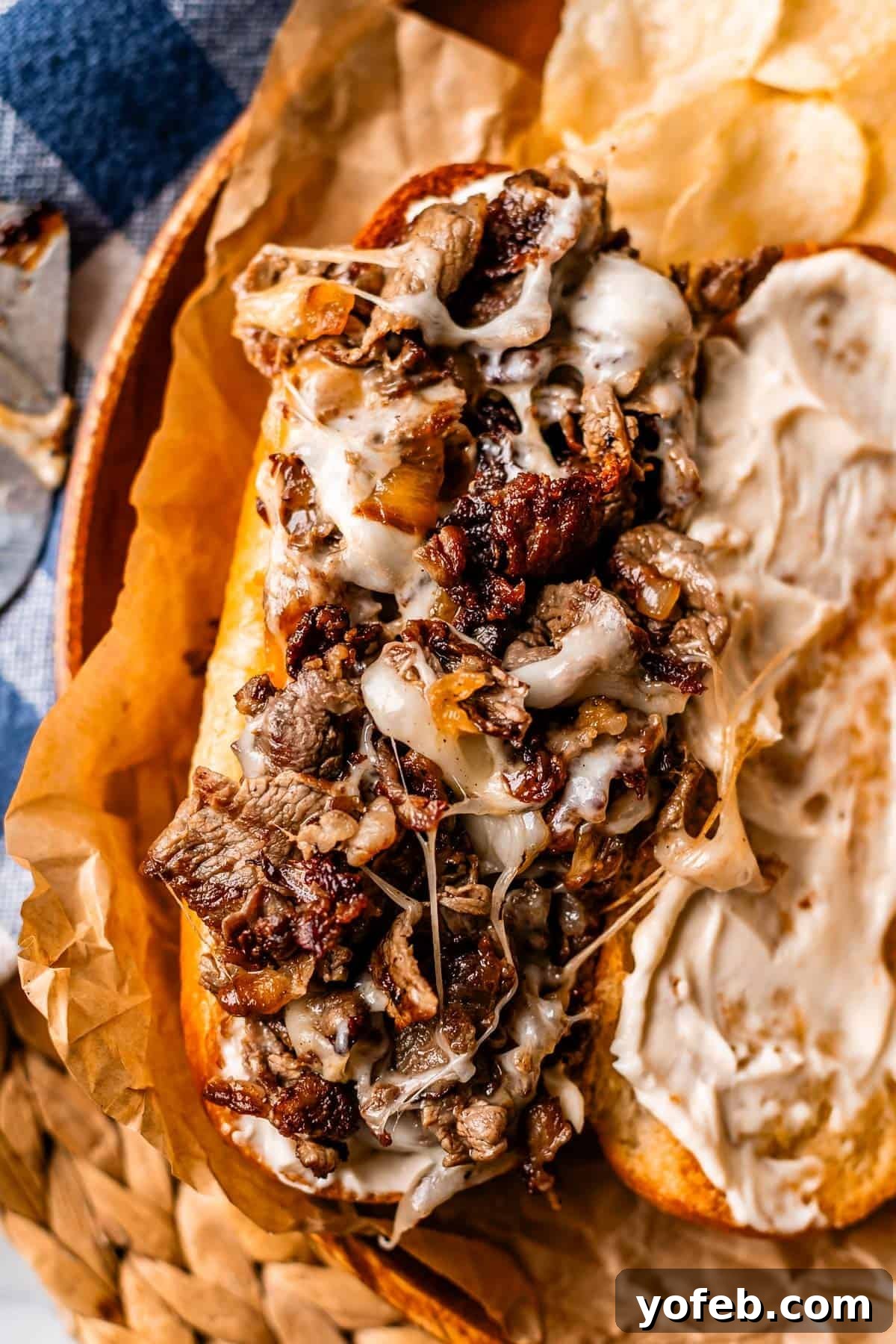 Authentic Philly Cheesesteak Recipe: How to Make It at Home 16 Close-up of a toasted sandwich filled with melted cheese and chopped, grilled beef, served on brown parchment paper with potato chips on the side. One half of the bread is spread with a creamy white sauce.