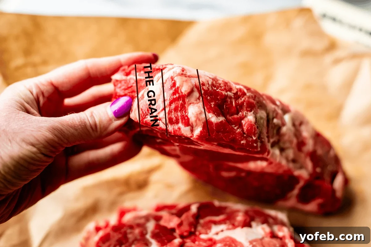 Authentic Philly Cheesesteak Recipe: How to Make It at Home 9 A hand with purple nail polish holds a raw cut of ribeye showing the direction of the grain, labeled with the words THE GRAIN and three lines for reference, over brown butcher paper.