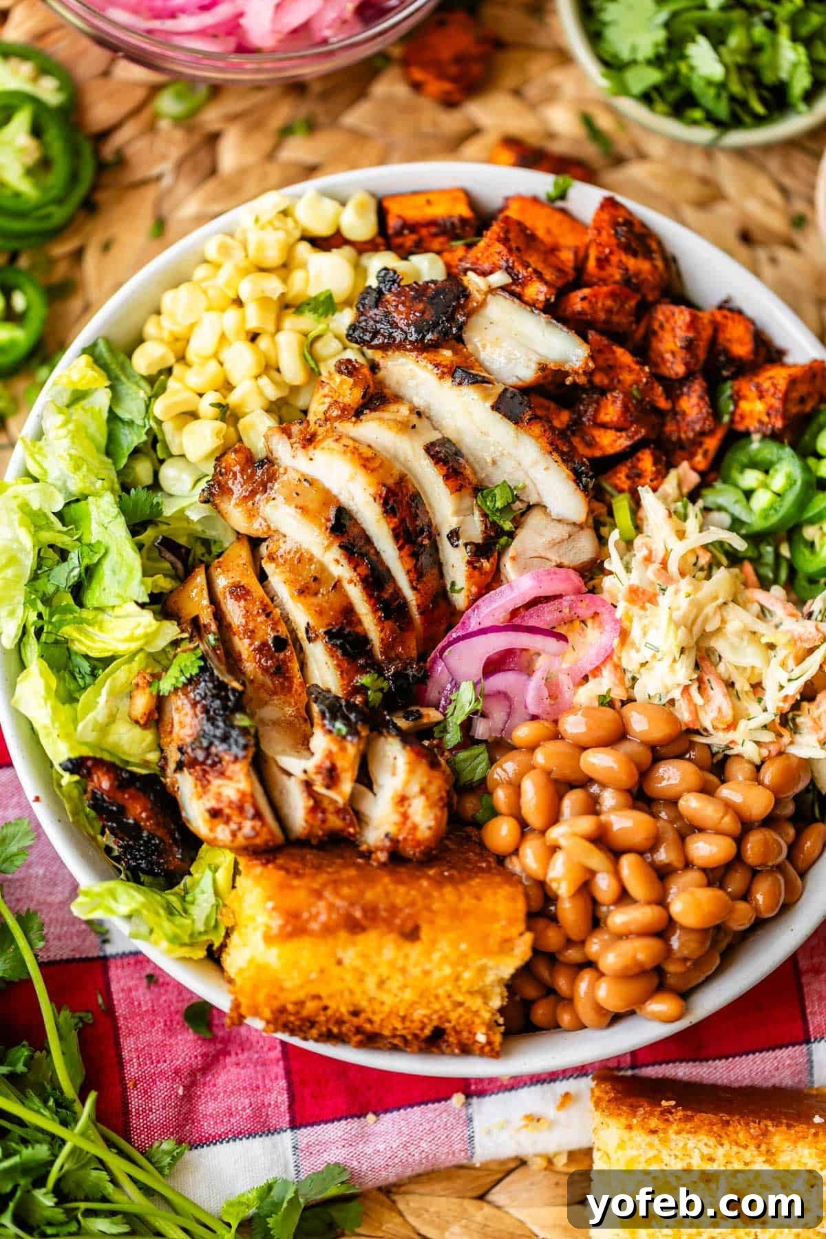 Smoky BBQ Chicken Rice Bowls with Grilled Veggies 9 A bowl filled with sliced grilled chicken, corn, lettuce, roasted sweet potatoes, coleslaw, pickled onions, pinto beans, jalapeños, and a piece of cornbread, arranged on a colorful picnic cloth.