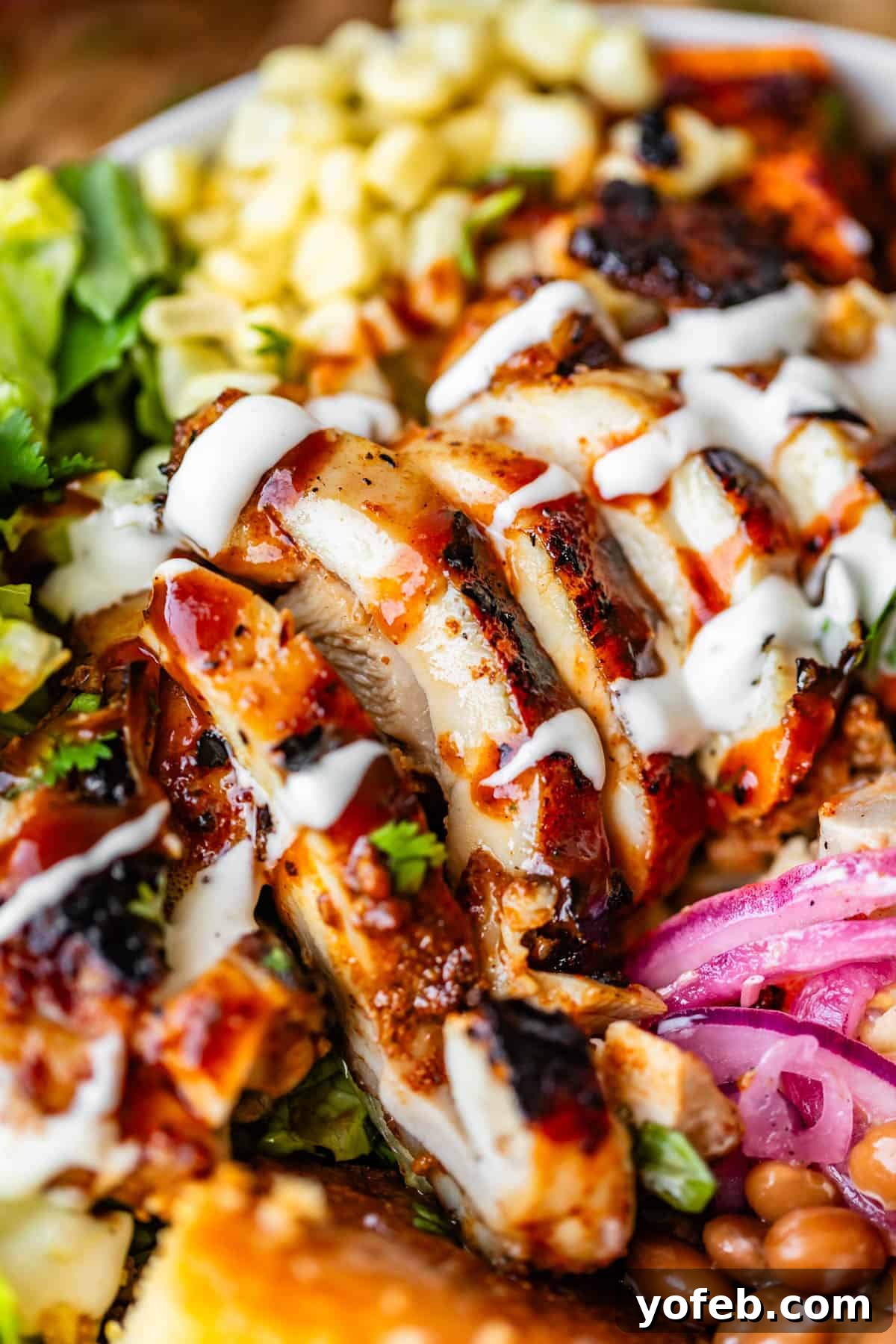 Smoky BBQ Chicken Rice Bowls with Grilled Veggies 10 Close-up of a salad bowl with sliced grilled chicken drizzled with barbecue sauce and ranch dressing, surrounded by corn, lettuce, beans, pickled onions, and a slice of cornbread.