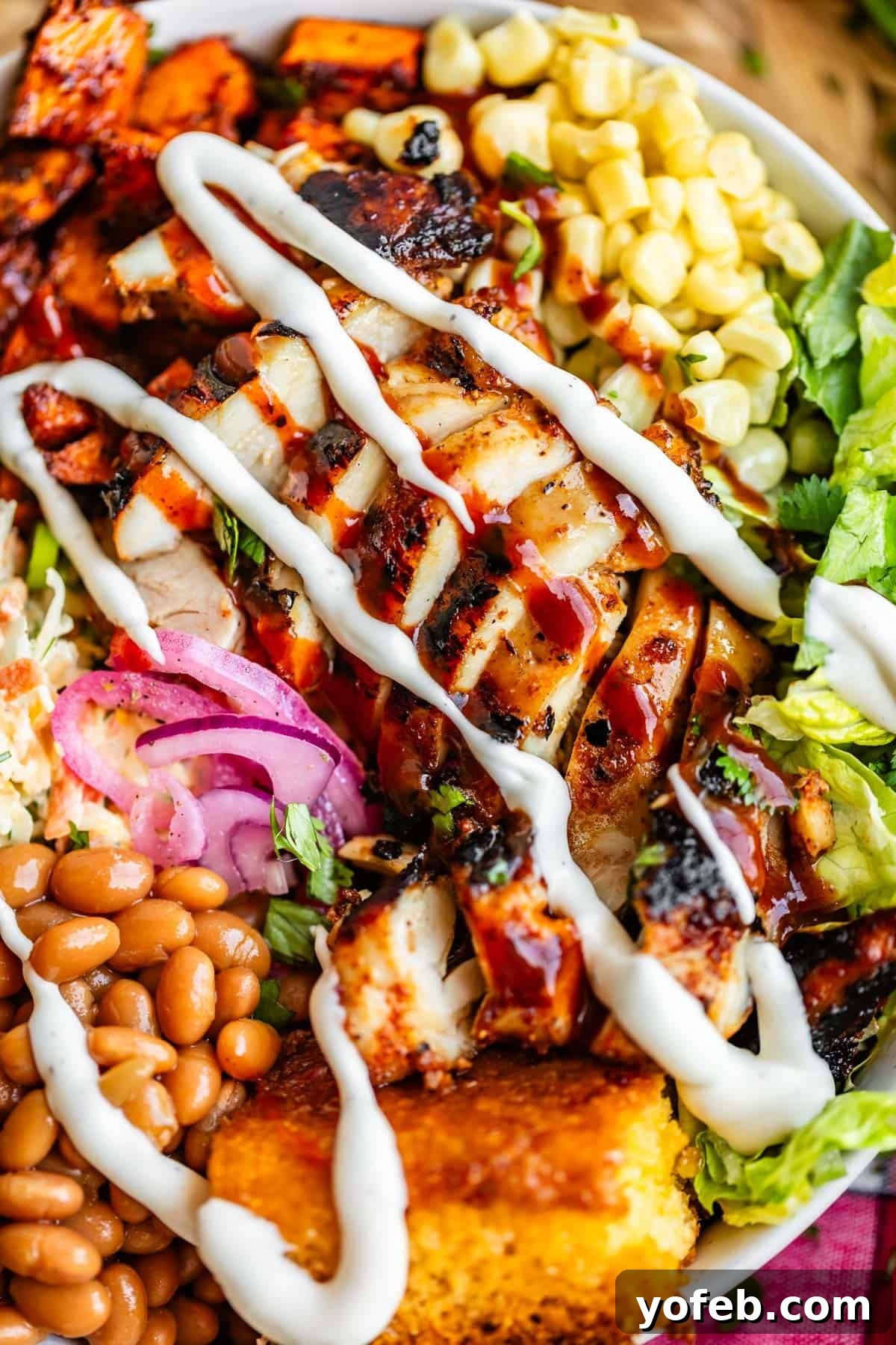 Smoky BBQ Chicken Rice Bowls with Grilled Veggies 11 A colorful plate with grilled chicken drizzled with white sauce, surrounded by baked beans, cornbread, corn, lettuce, pickled onions, coleslaw, and roasted sweet potatoes.