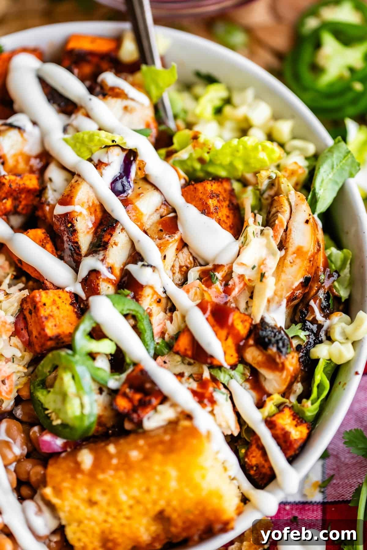 Smoky BBQ Chicken Rice Bowls with Grilled Veggies 3 A close-up of a colorful salad bowl with grilled chicken, spiced sweet potatoes, jalapeño slices, beans, shredded lettuce, cornbread, and a drizzle of creamy dressing.