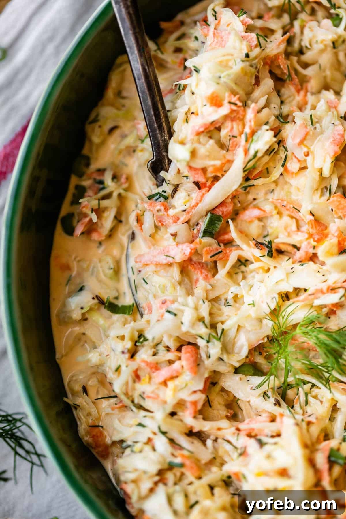 Classic Creamy Coleslaw Recipe with Tangy Dressing 13 Close-up of creamy coleslaw with shredded cabbage, carrots, fresh dill, and a mayonnaise-based dressing in a green bowl, with a serving spoon partially visible.
