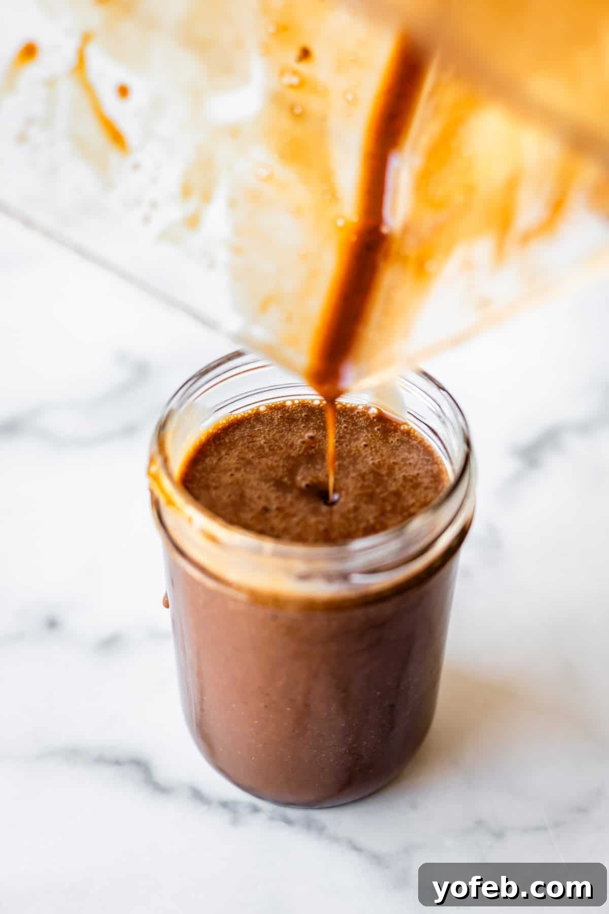 Creamy Balsamic Vinaigrette for Salads 7 A thick, dark brown liquid is being poured from a blender into a glass jar on a white marble surface. Some of the liquid is visible inside the blender, and the jar is nearly full.