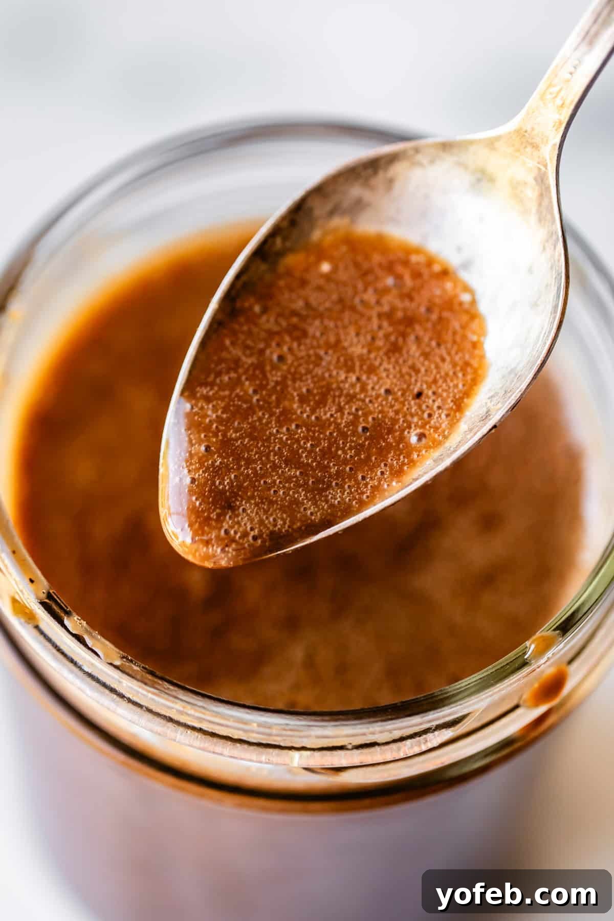 Creamy Balsamic Vinaigrette for Salads 9 A close-up of a spoon dipped into a jar of thick, brown sauce or dressing, with the spoon held above the jar showing its smooth, glossy texture.