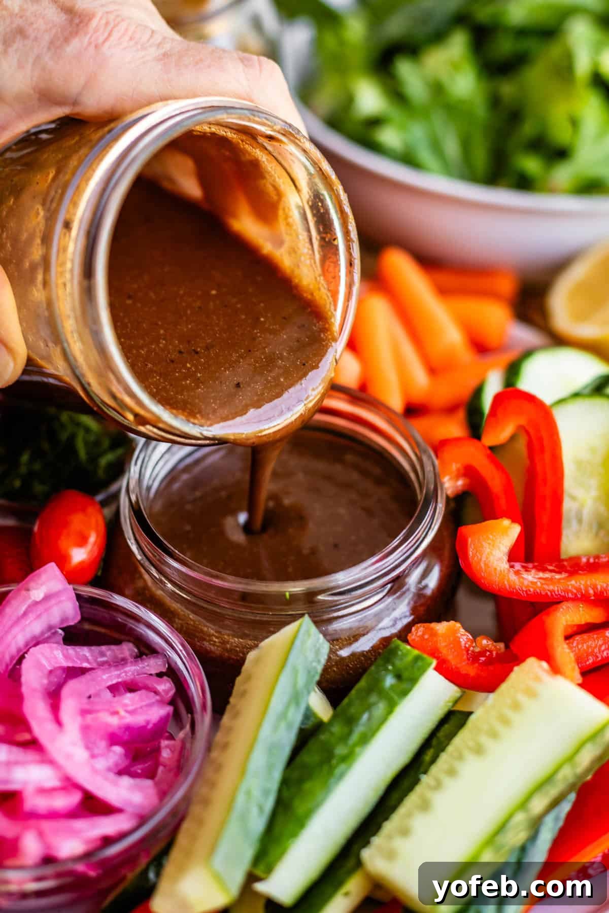 Creamy Balsamic Vinaigrette for Salads 3 A hand pours dark brown salad dressing from a jar into a small bowl, surrounded by fresh vegetables including cucumbers, red bell peppers, carrots, lettuce, and pickled onions.