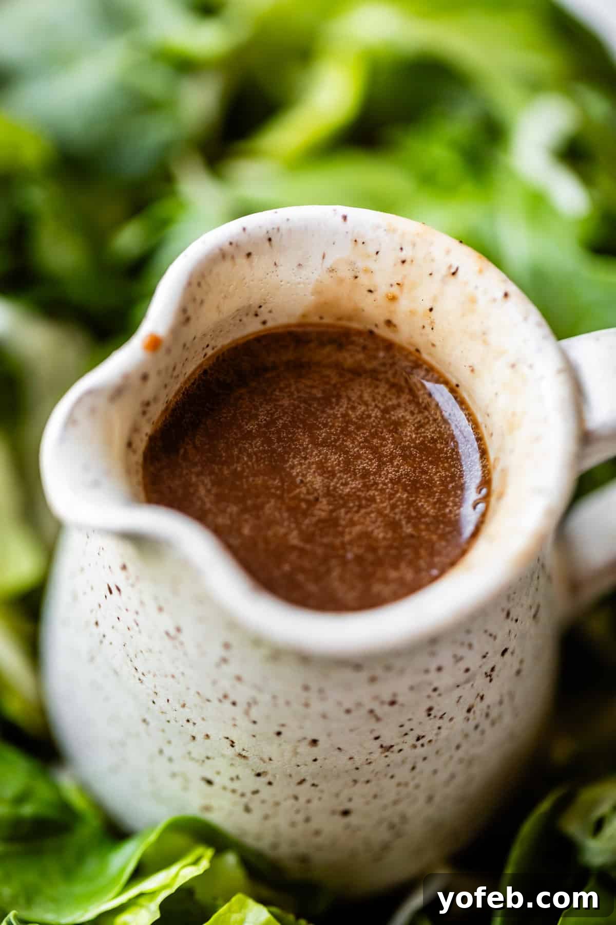 Creamy Balsamic Vinaigrette for Salads 4 A close-up of a small ceramic pitcher filled with brown vinaigrette dressing, surrounded by fresh green leafy vegetables.